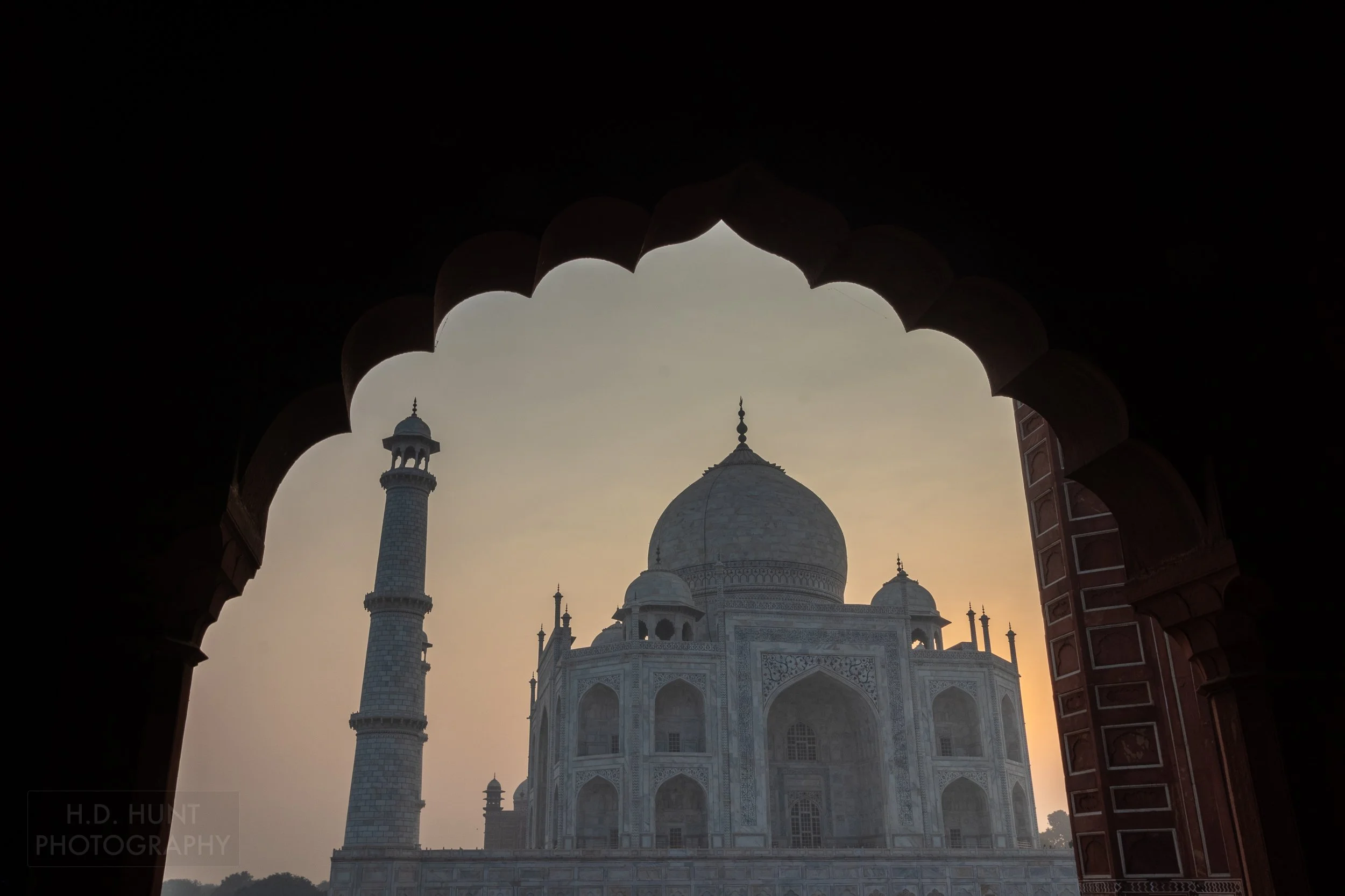 An orange sky is seen behind the Taj Mahal, Agra, India, as viewed through an archway with circular edges.