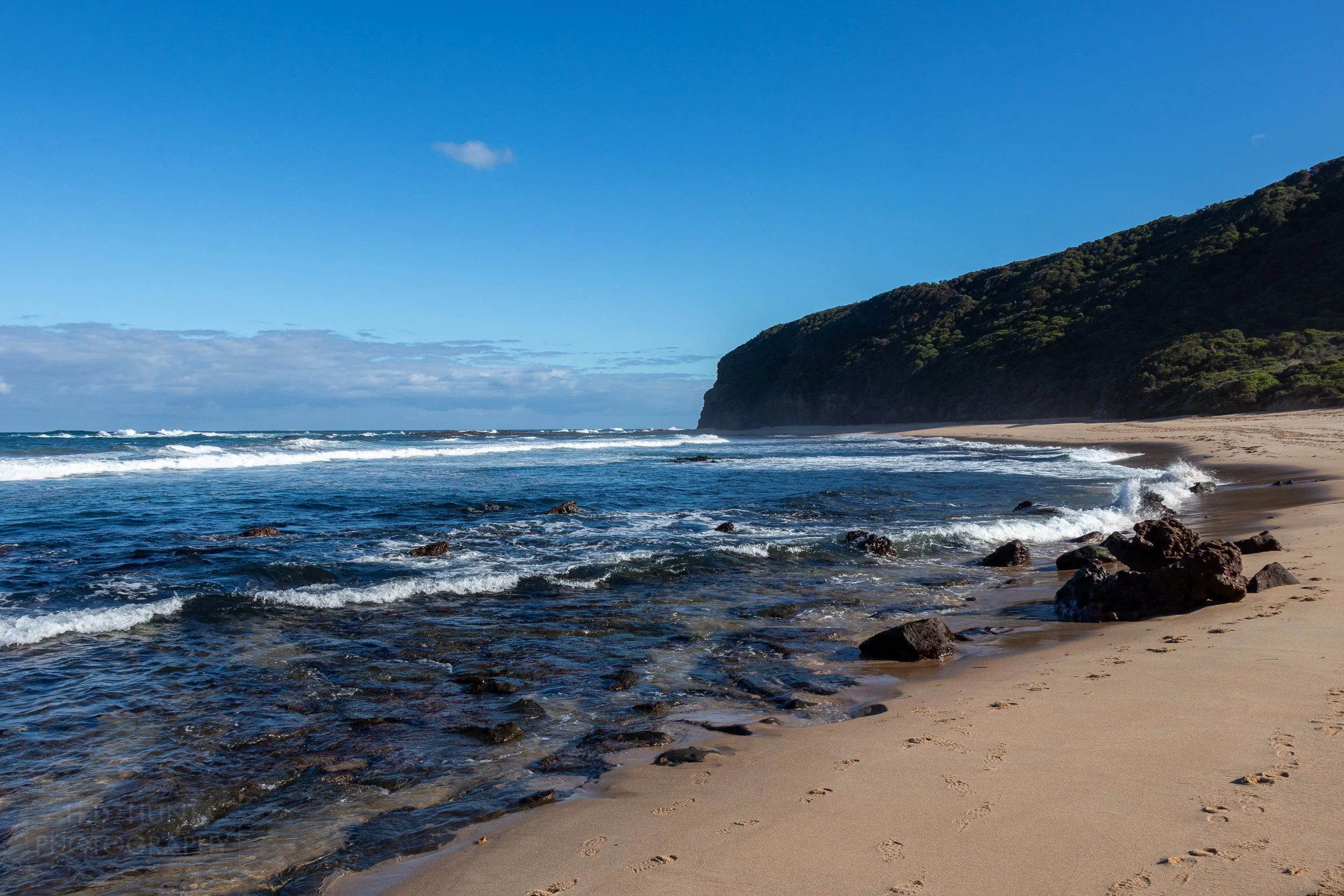 The sea and large rocks meet brown sands beneath cliffs at Wreck Beach on The Great Ocean Walk, Victoria, Australia.