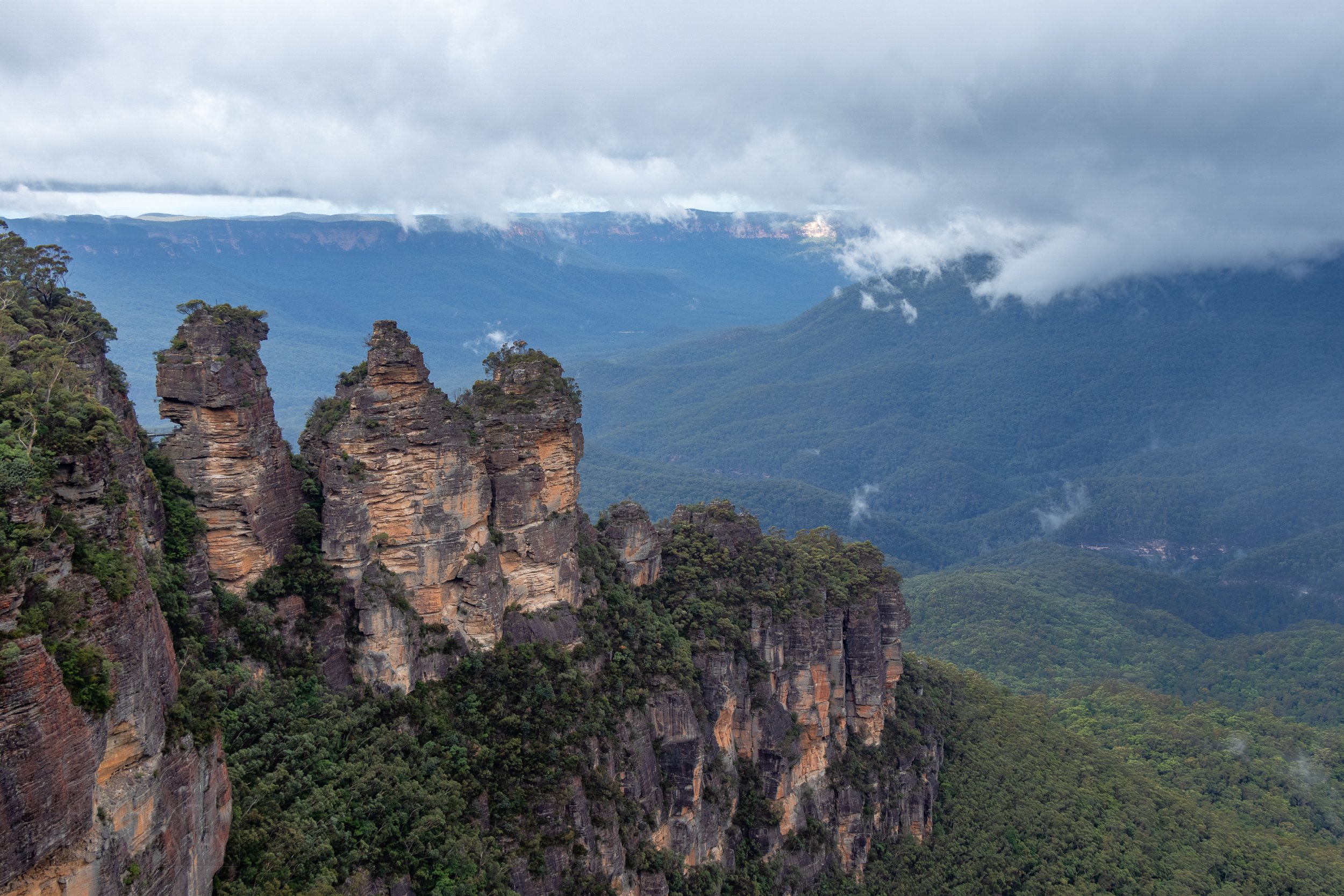 The Three Sisters sandstone column rock formation stands in front of a large foggy valley, Katoomba, Australia.