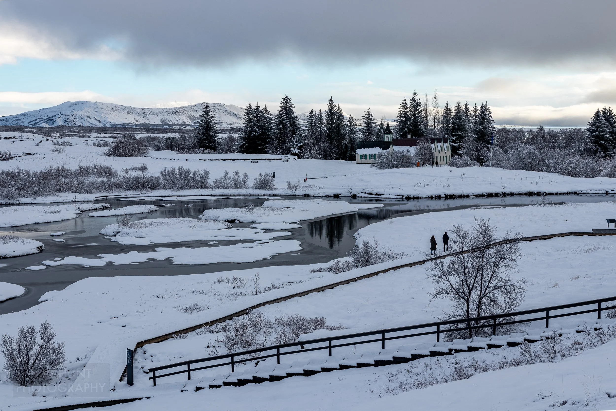Two visitors walk beside a river on the other side of which is found a yellow building at Þingvellir, Iceland.