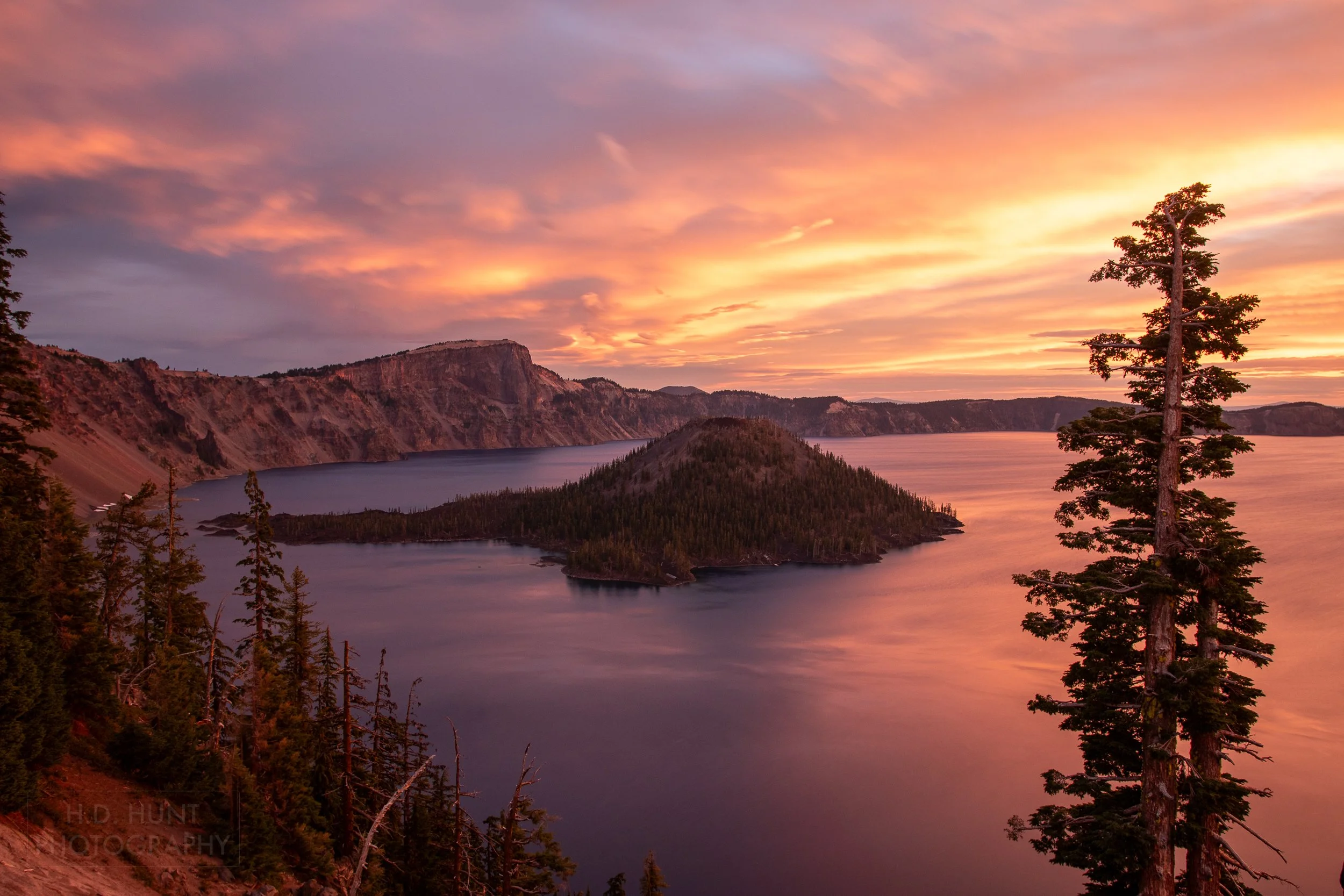 The sunrise creates a pink, orange, and yellow sky above the blue waters of Crater Lake and Wizard Island, Crater Lake National Park, Oregon, United States.