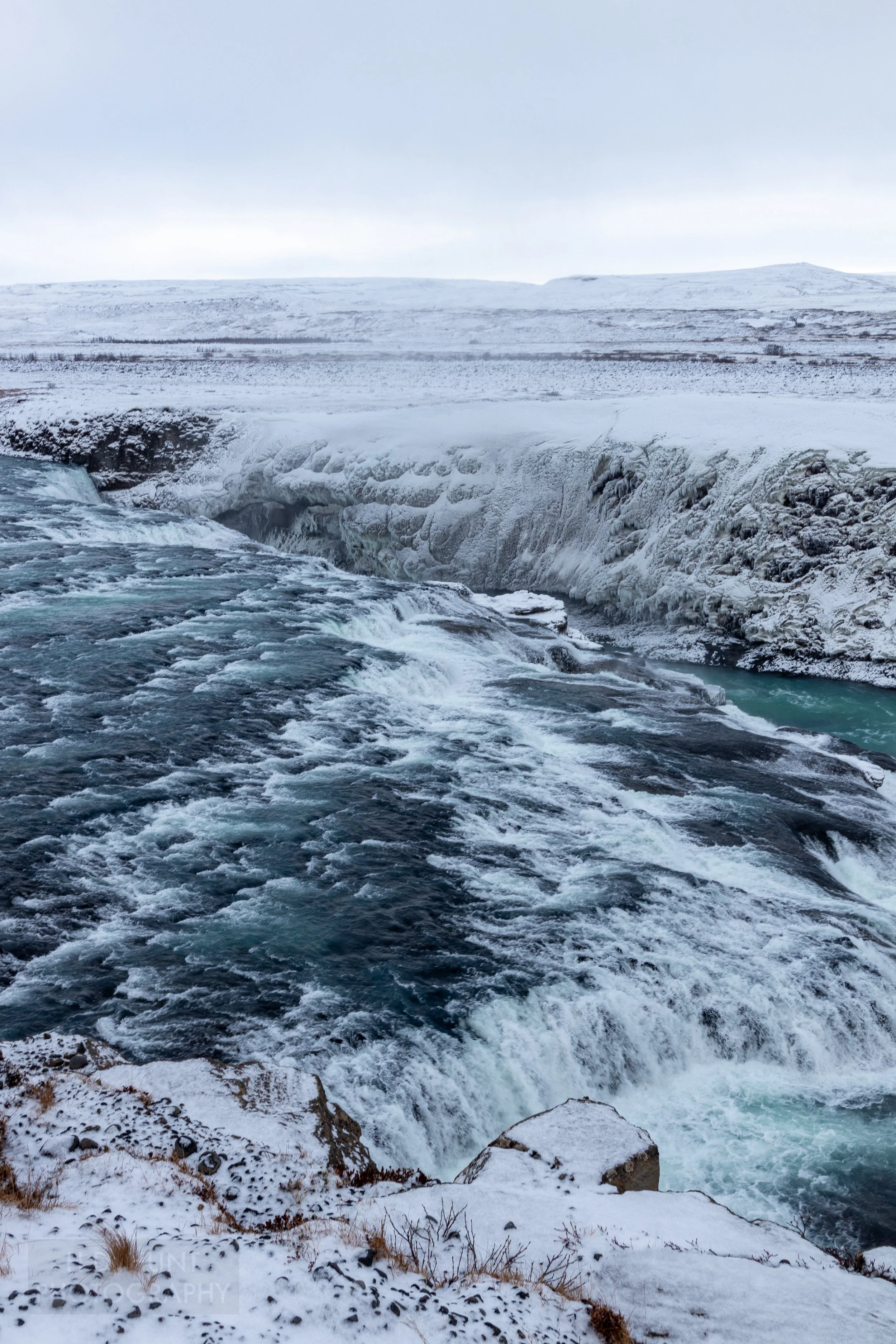 A waterfall, Gullfoss, features dark blue water passing over several tiers of falls surrounded by snow-covered rock cliffs, Iceland.