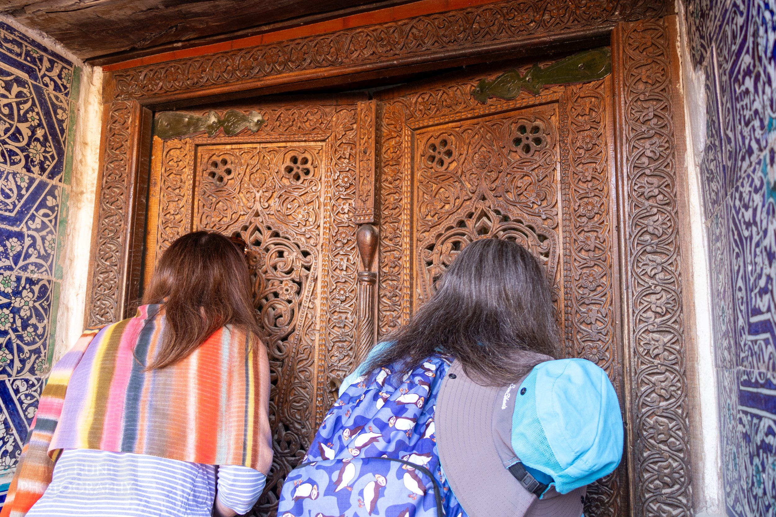 Two visitors look through a large wooden door in the Reception Courtyard of the Kuhna Ark, Khiva, Uzbekistan.