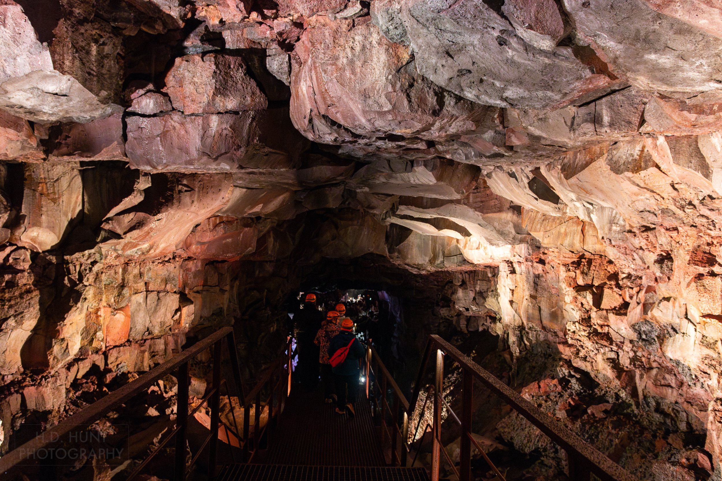 Visitors walk along a metal walkway beneath a stone ceiling within the lava tube Raufarhólshellir, Iceland.