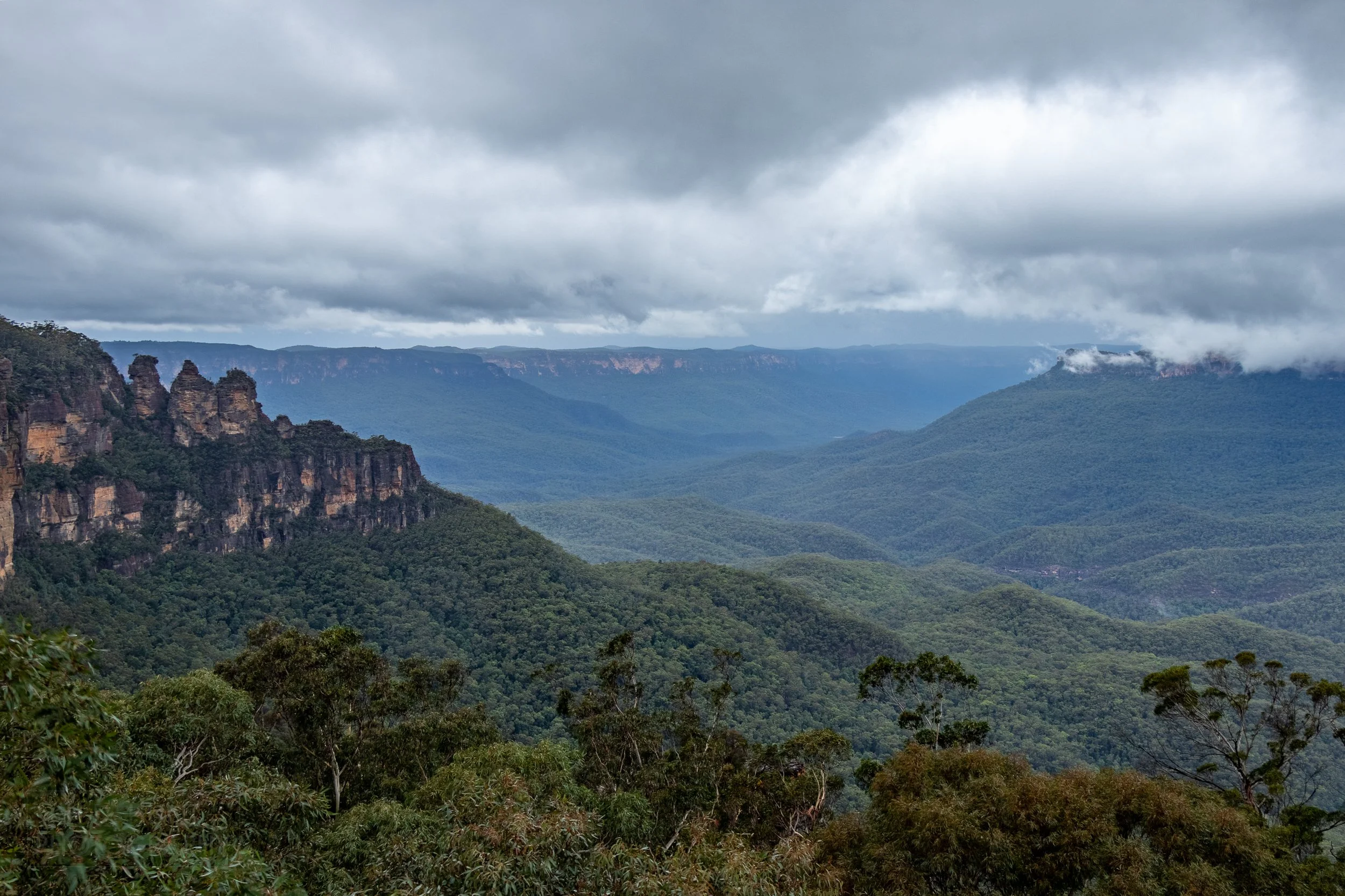 The Three Sisters sandstone column rock formation stands in front of a large foggy valley, Katoomba, Australia.