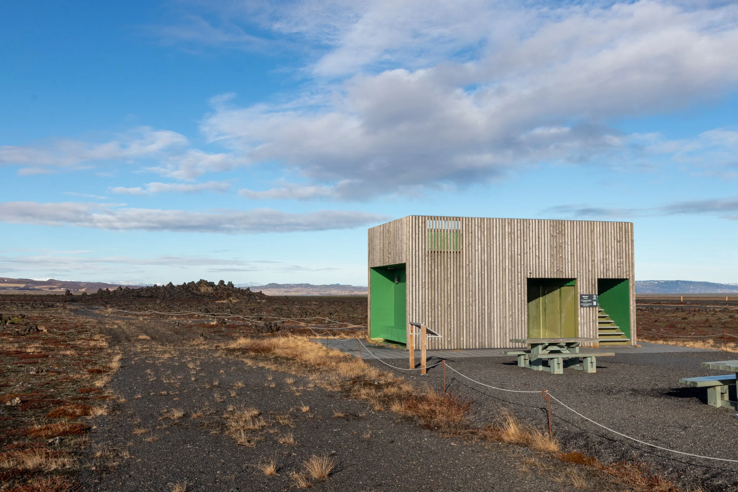 A wood-paneled green toilet block stands in the foreground of a lava field containing cairns of basalt, Laufskálavarða, Iceland.