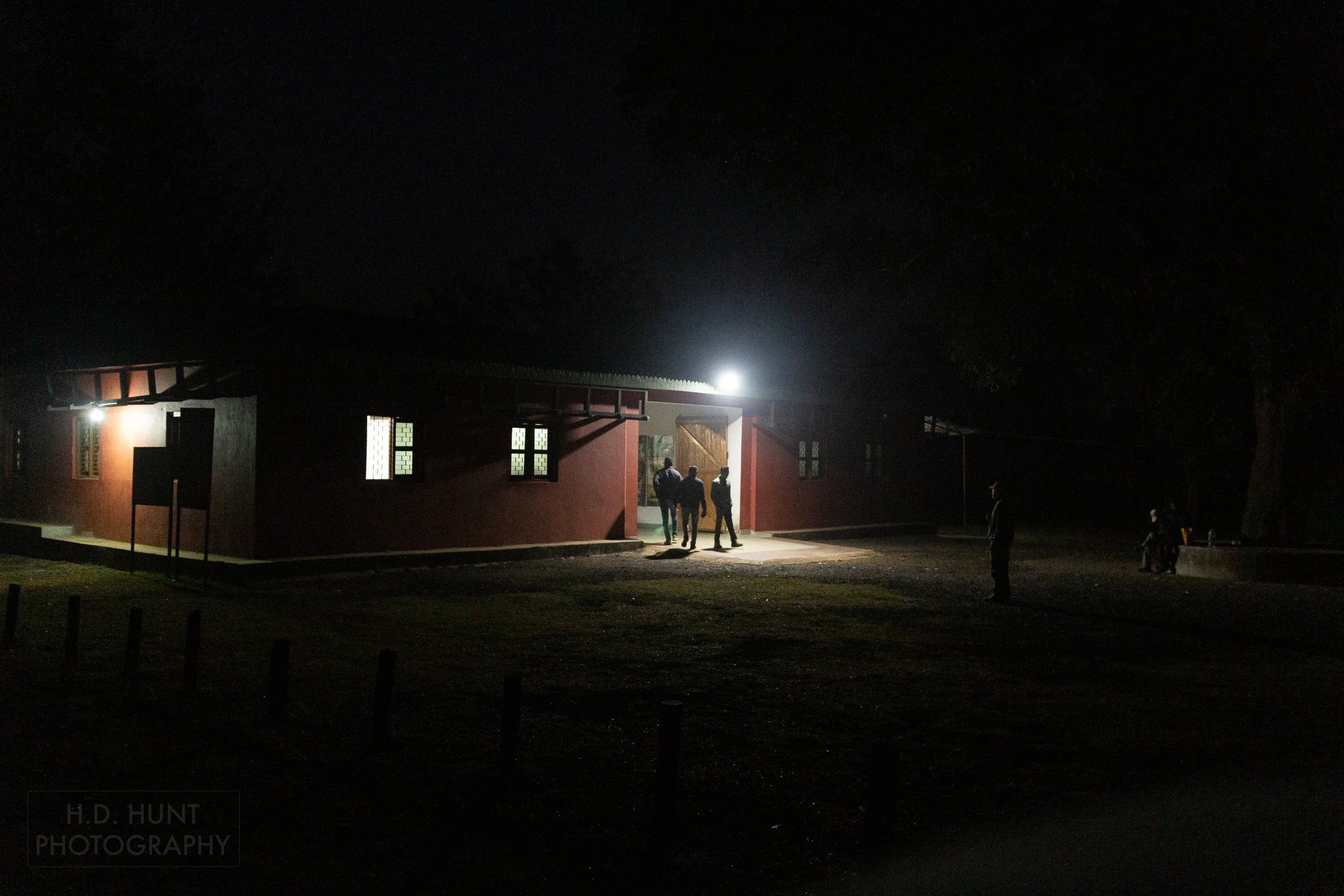 Three guides emerge from a red building surrounded by the pre-dawn darkness, Kanha Tiger Reserve, India.