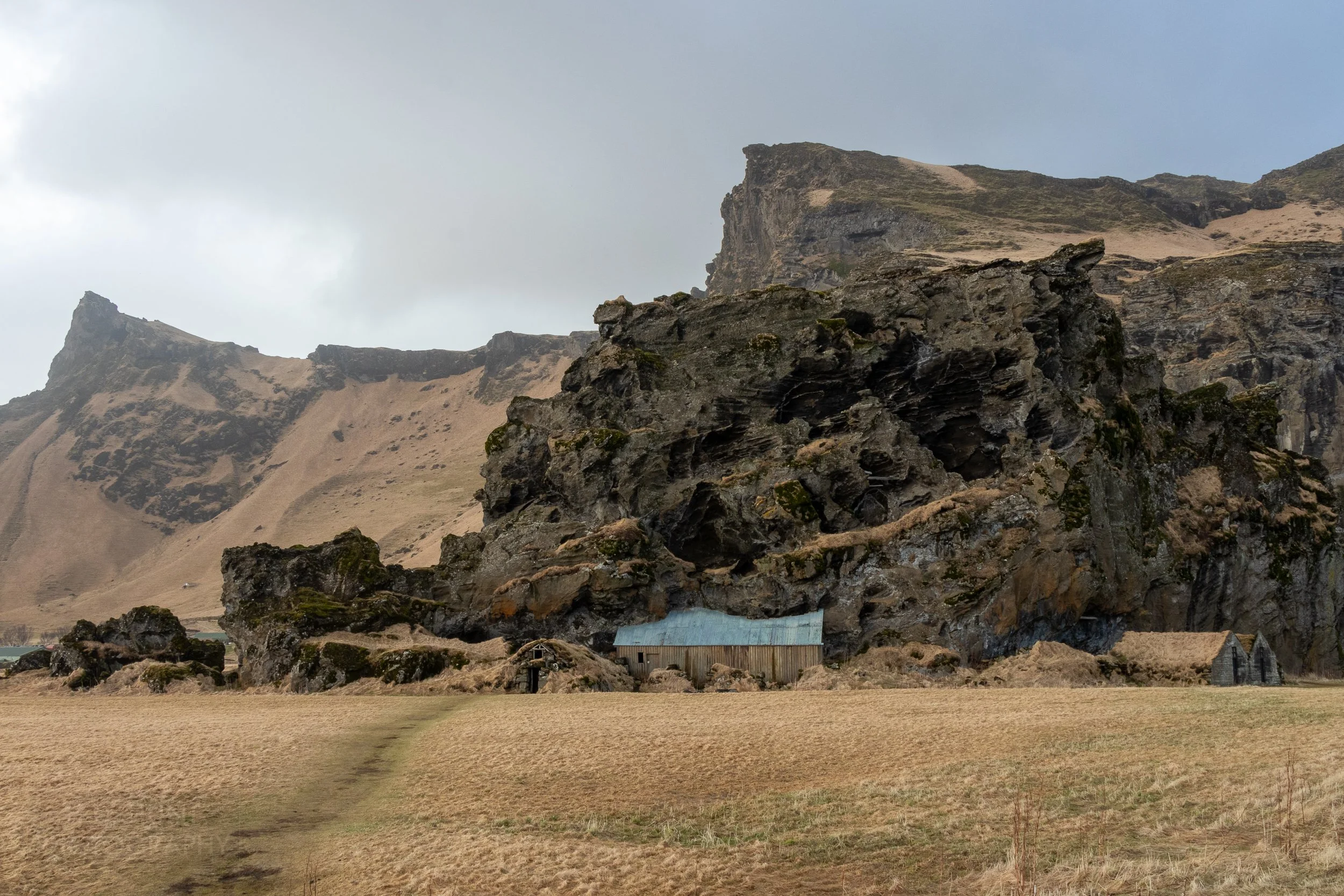 Several sheds are nestled underneath a large black rock in a brown grass field, Drangurinn Rock, Iceland.