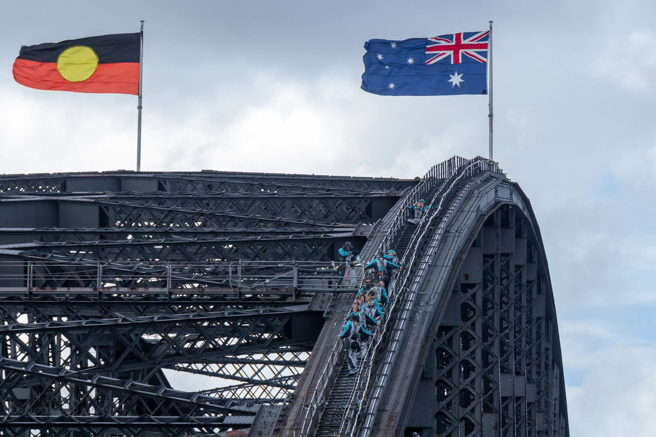 The dark-colored steel of the Sydney Harbor Bridge rises while light blue-clad visitors climb the bridge's outer supports beneath large Australian and Aboriginal flags, Sydney, Australia.