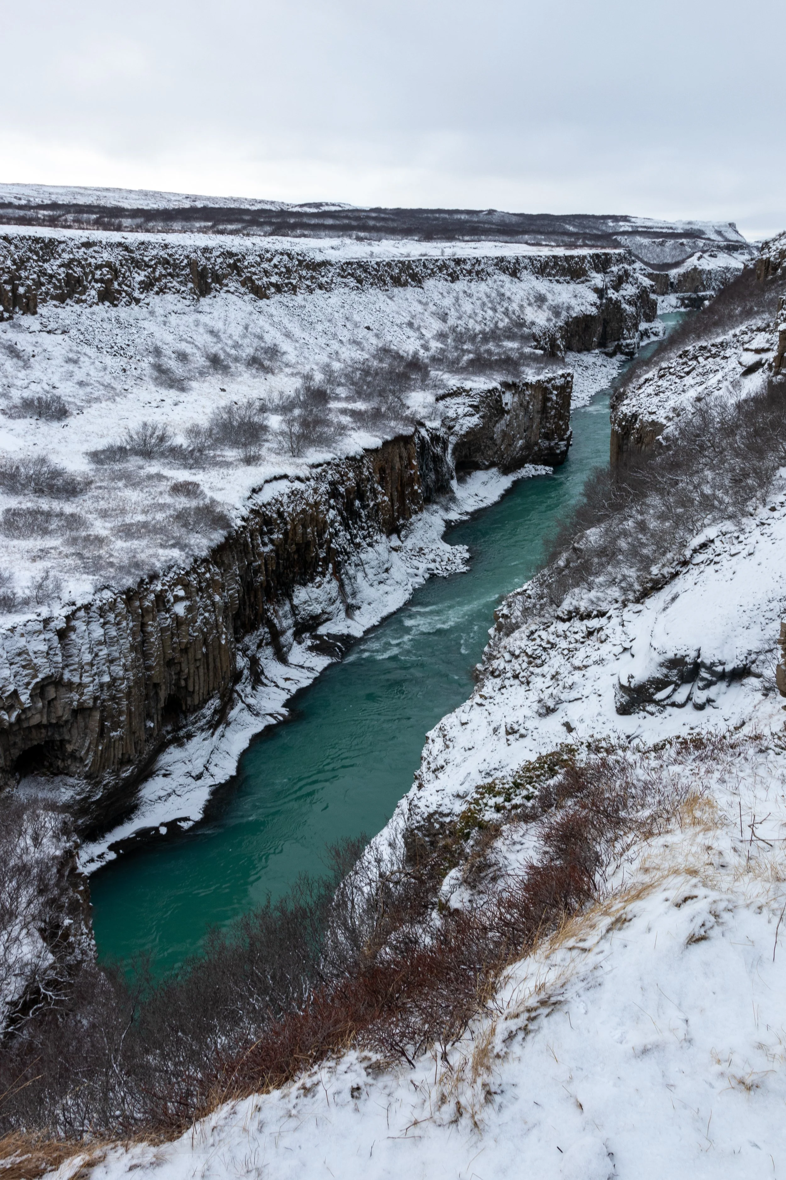 Dark blue water passes over several tiers of falls surrounded by snow-covered rock cliffs, Iceland.