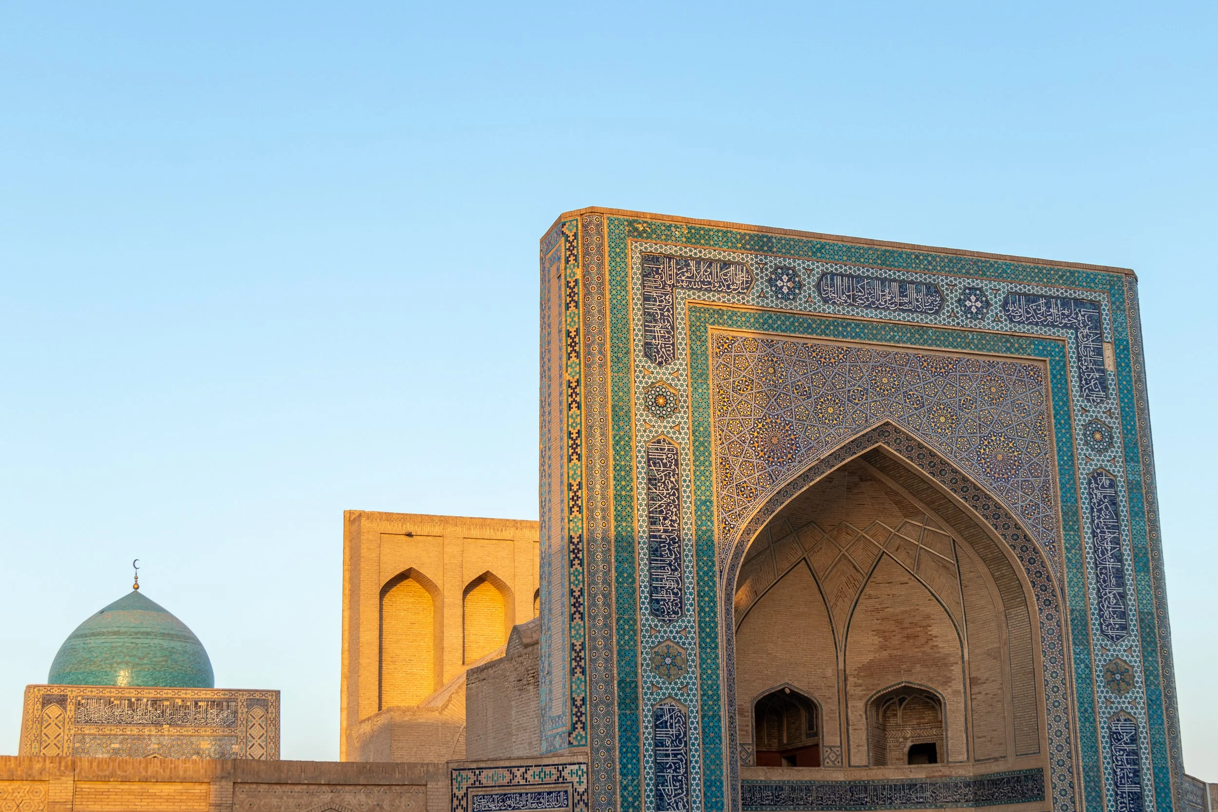 The large blue dome and tiled facade of the Kalan Mosque is illuminated by the sunrise, Bukhara, Uzbekistan.