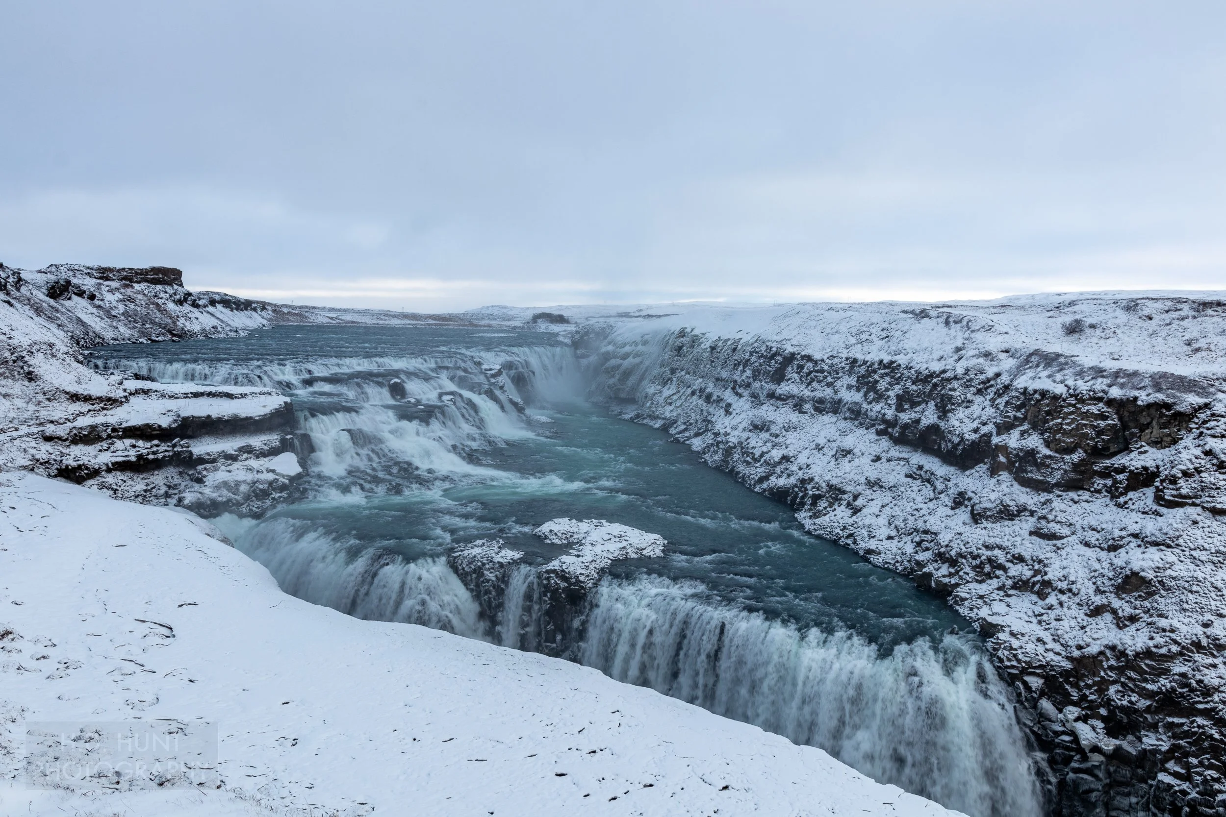 A waterfall, Gullfoss, features dark blue water passing over several tiers of falls surrounded by snow-covered rock cliffs, Iceland.