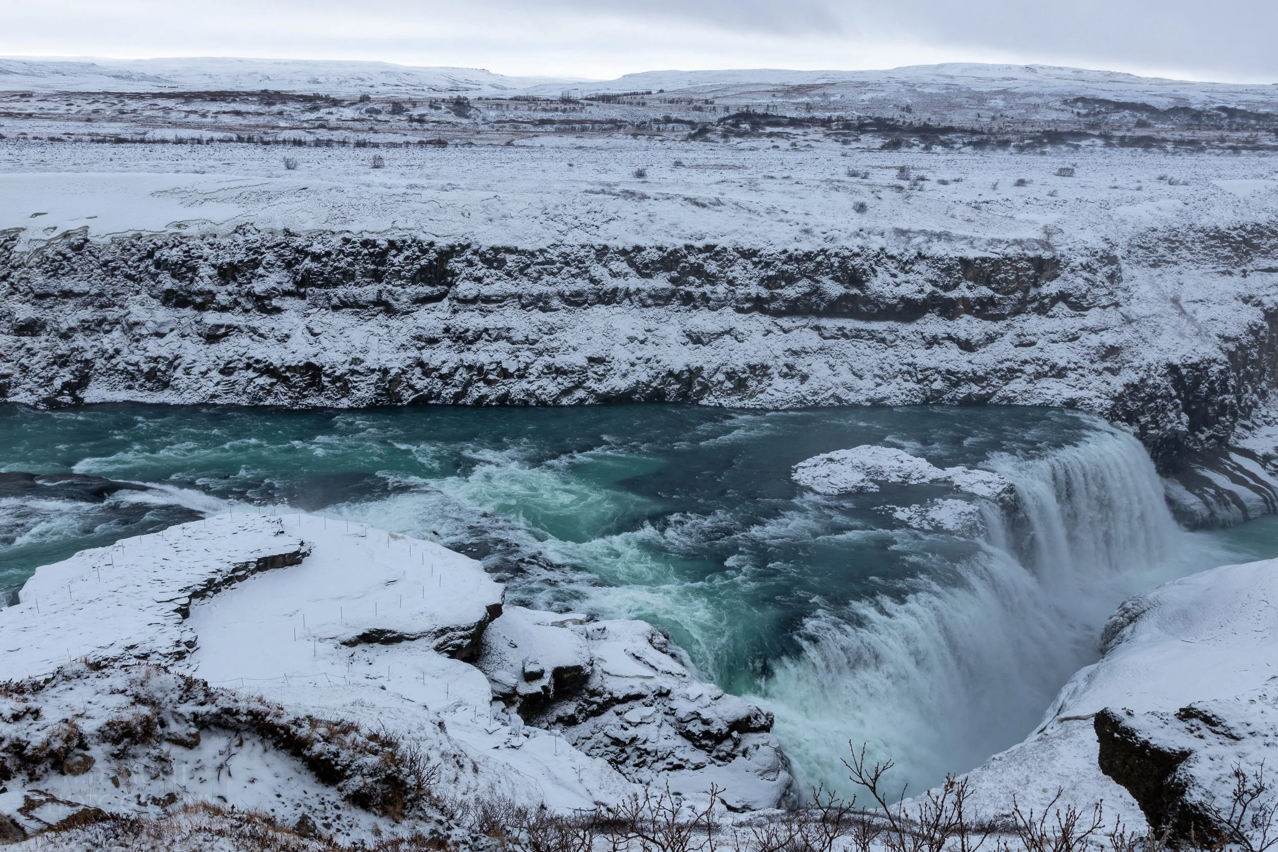 A waterfall, Gullfoss, features dark blue water passing over several tiers of falls surrounded by snow-covered rock cliffs, Iceland.