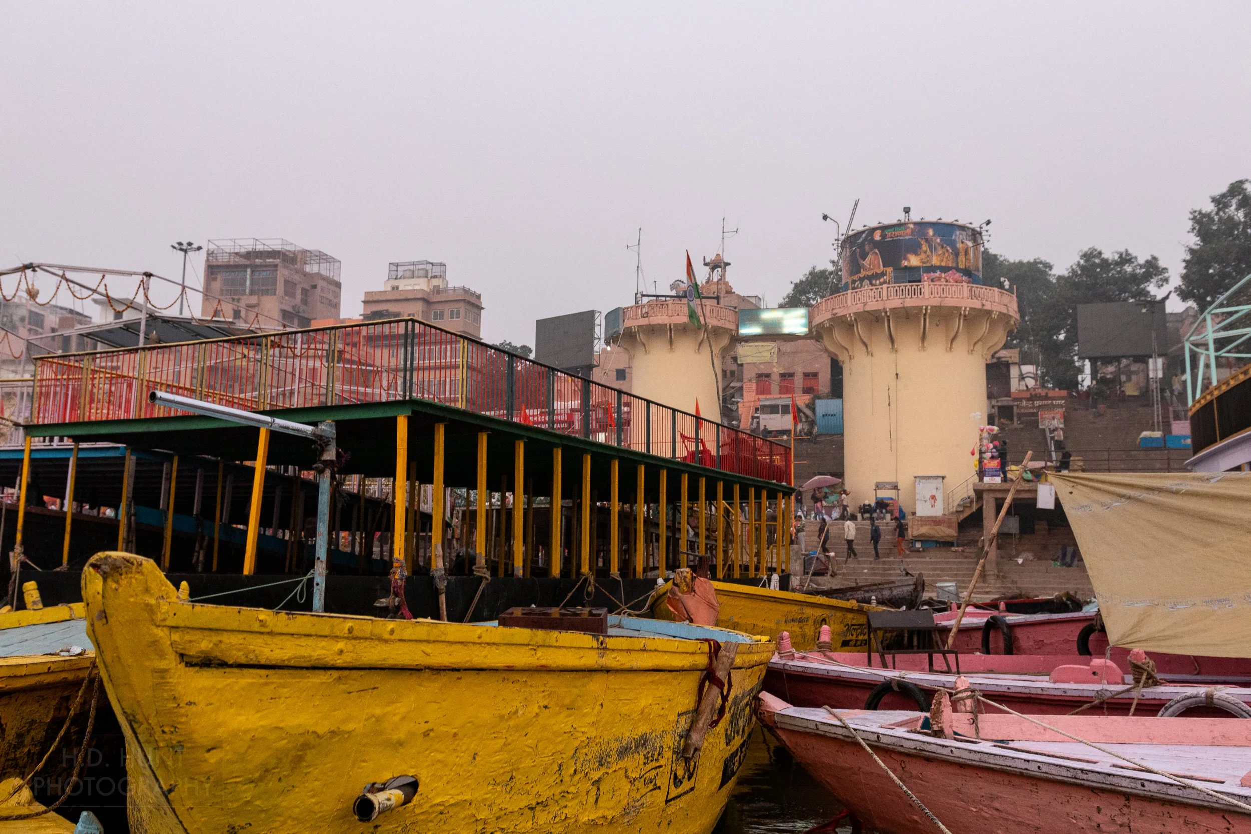 Two large yellow boats and two small pink boats are moored besides a tiered balcony, Varanasi, India.