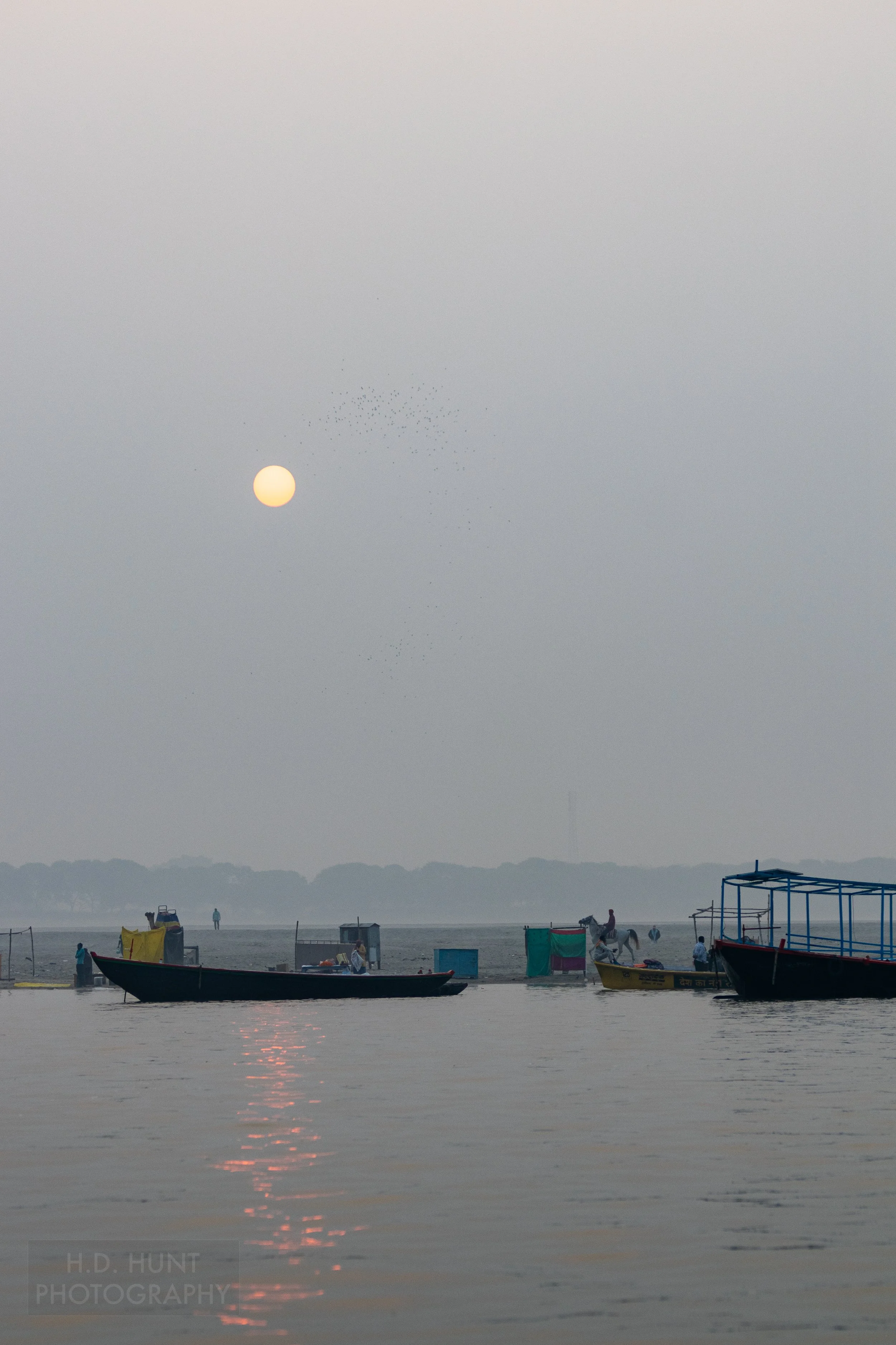 The sun rises over two boats and a man riding a horse, Varanasi, India.