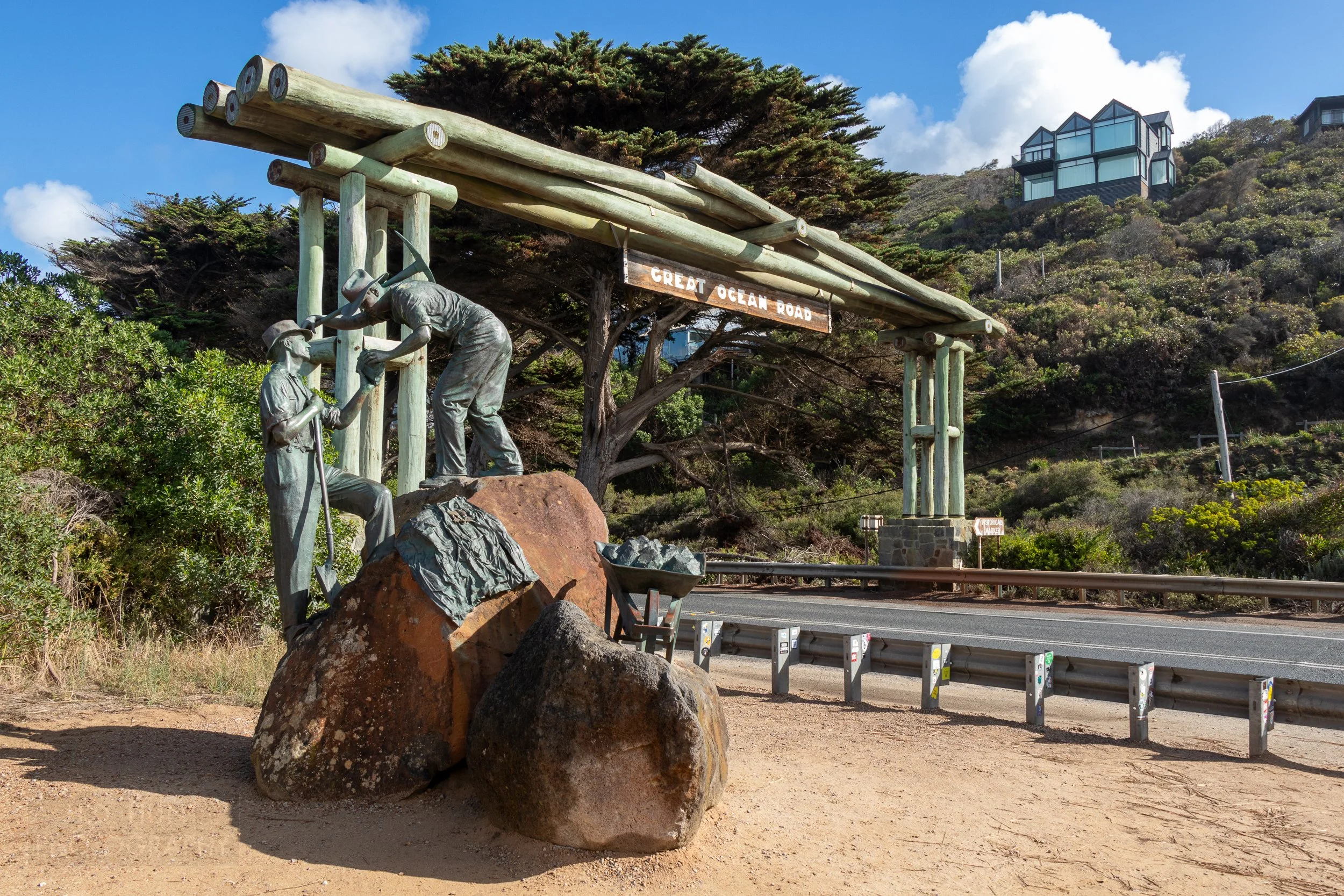 A metal statue of two male laborers with pickaxes and shovels sits atop a brown rock near an arch over a roadway with a sign reading "Great Ocean Road", Victoria, Australia.