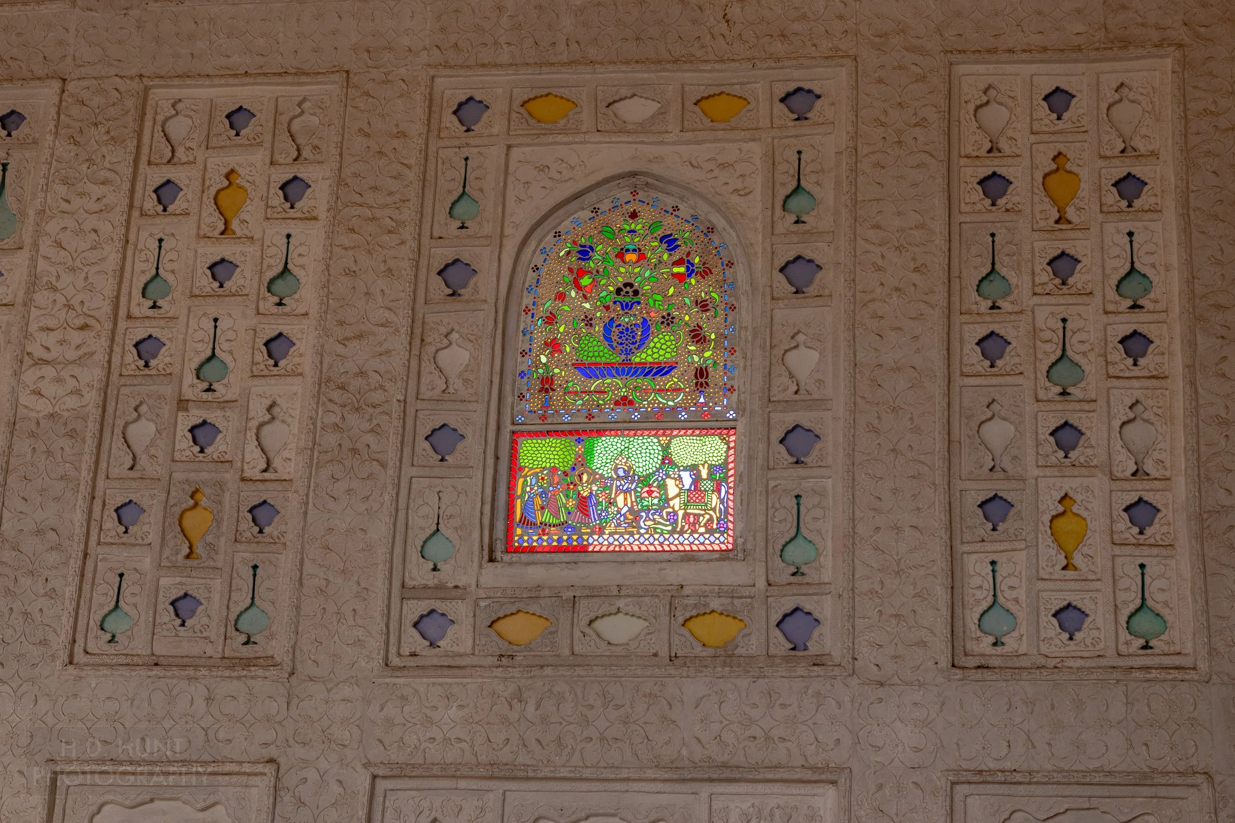 A stained glass window containing multiple small pieces is set in a wall with intricate carvings, Amber Fort, Amer, India.