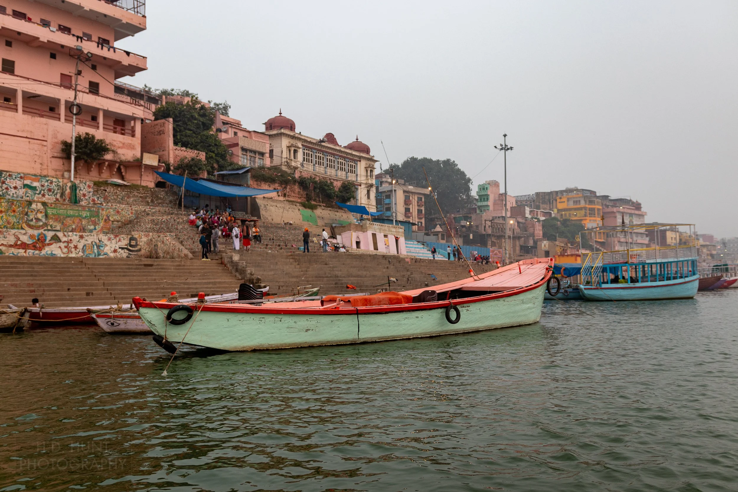 Boats are tied up along the Ganges River in front of multiple brightly-colored buildings, Varanasi, India.