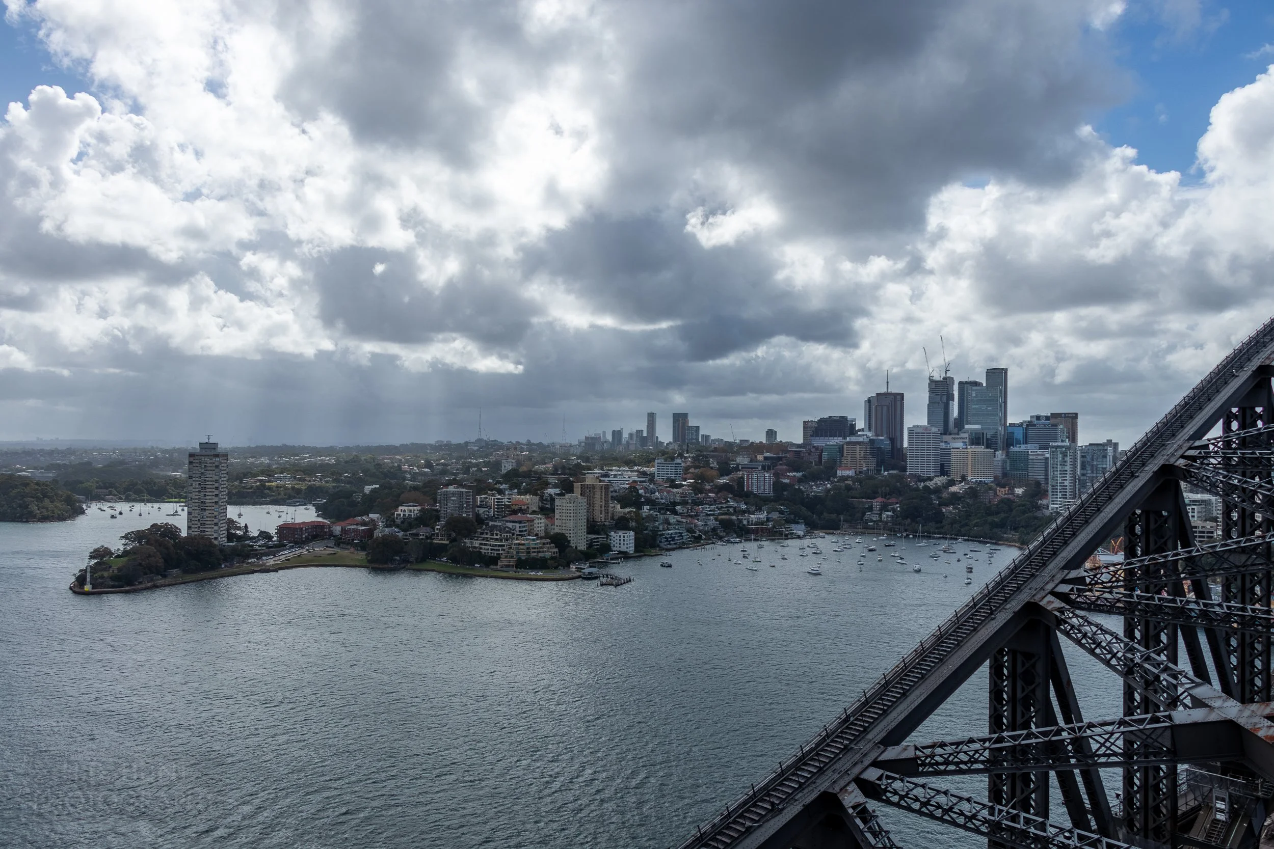 Light peaks through clouds to shine on a skyscraper-filled neighborhood behind the black steel of the Sydney Harbour Bridge, Sydney, Australia.