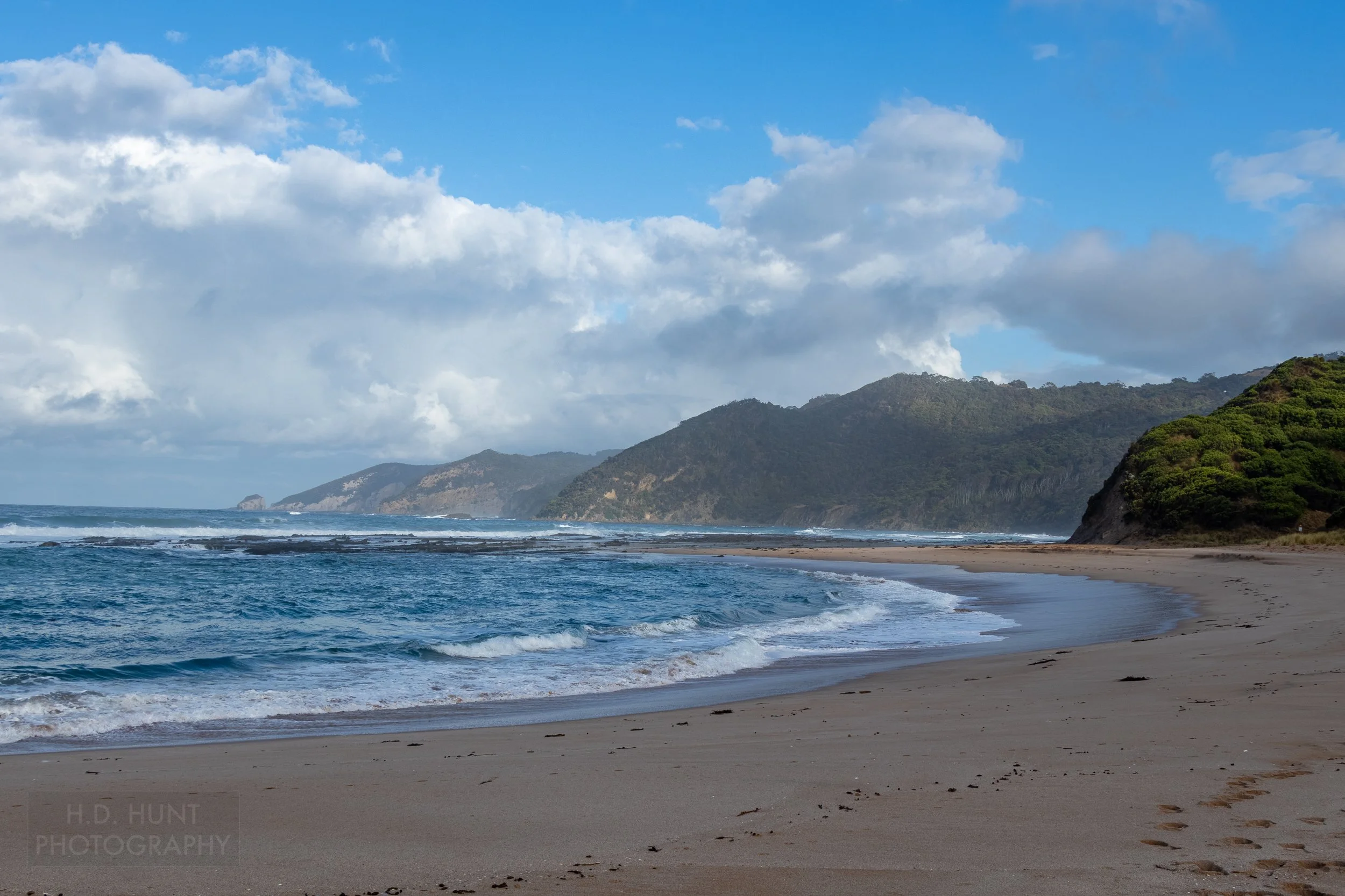A wide beach gives way to the sea beneath green-covered cliffs along The Great Ocean Walk, Victoria, Australia.