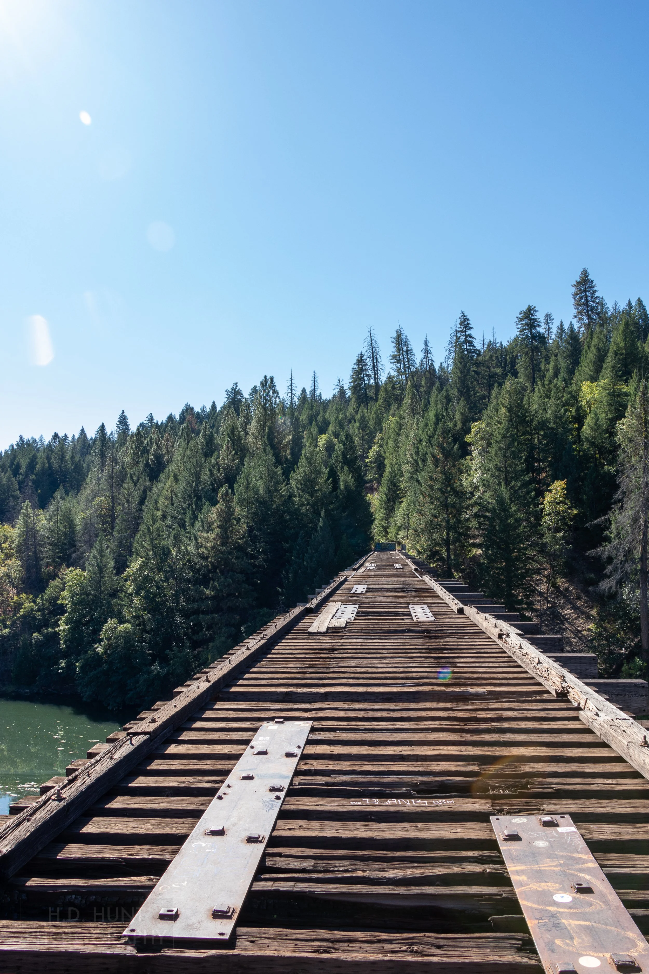 A railroad trestle bridge with brown timbers but no tracks spans a green river before disappearing behind trees, California, United States.