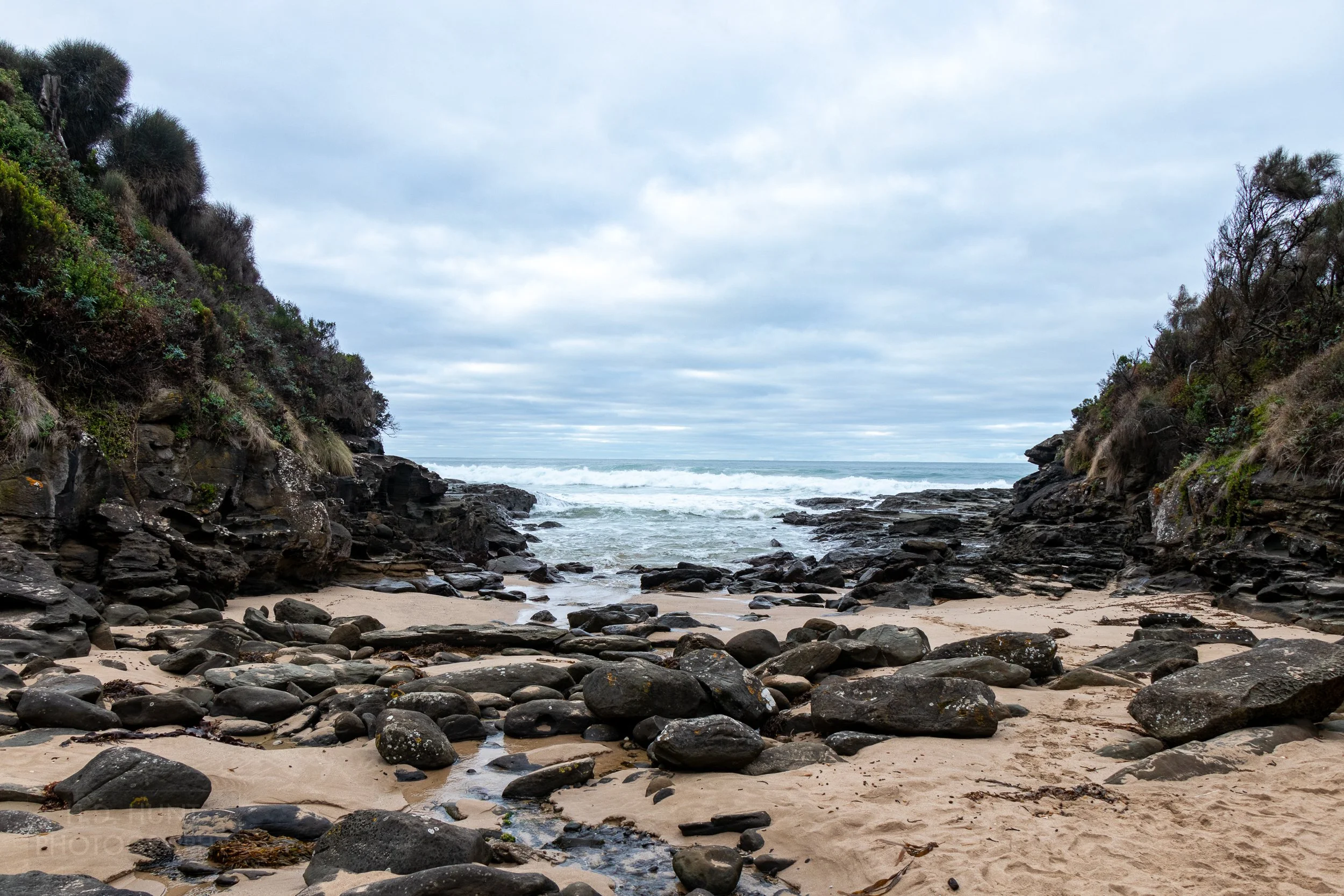 A small beach inside a cove filled with rocks is seen between two cliffs along The Great Ocean Walk, Victoria, Australia.