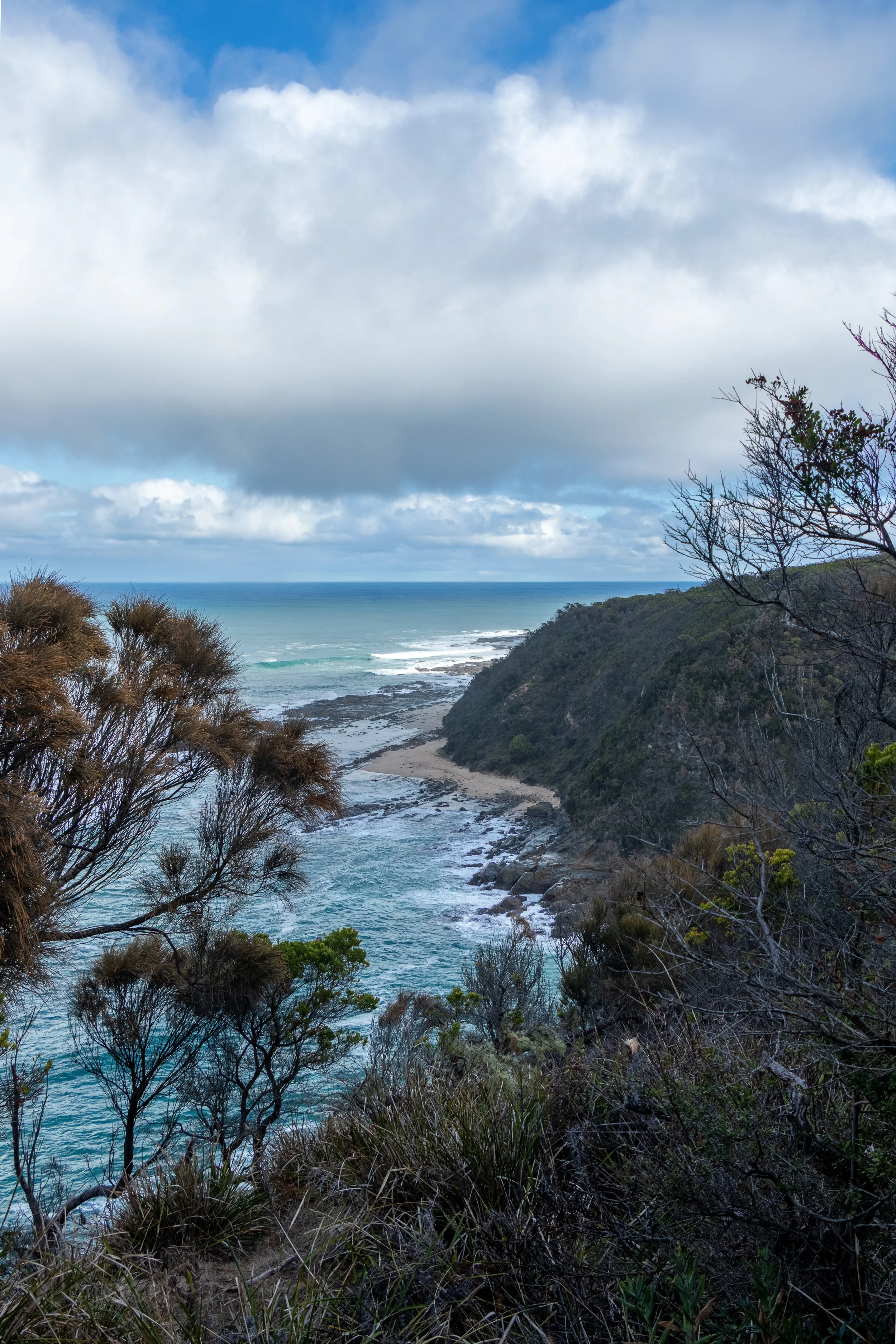 As seen from a cliff, the ocean meets a small beach and large boulders beneath a green plant-covered cliff along The Great Ocean Walk, Victoria, Australia.