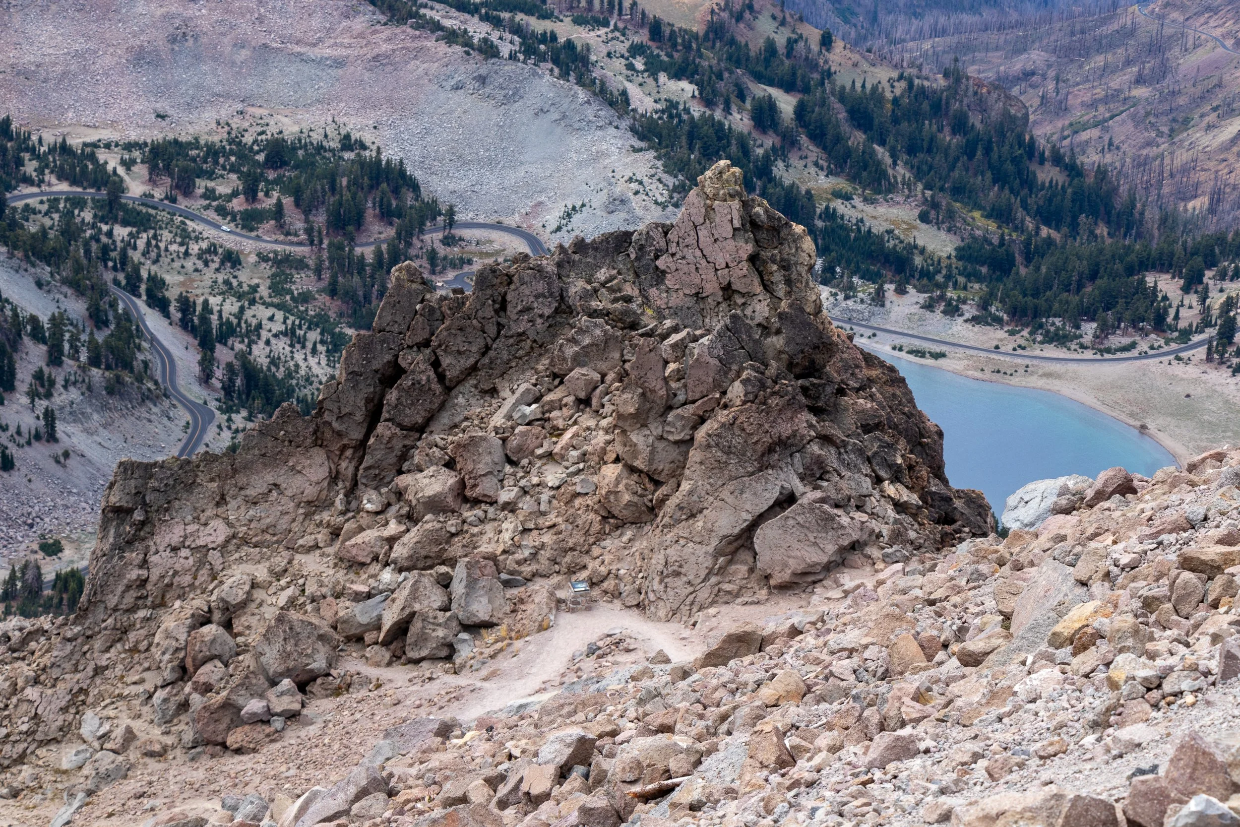 A crag pokes out from the earth in the foreground, while a roadway and blue lake can be seen in the background, Lassen Peak, Lassen Volcanic National Park, California, United States.