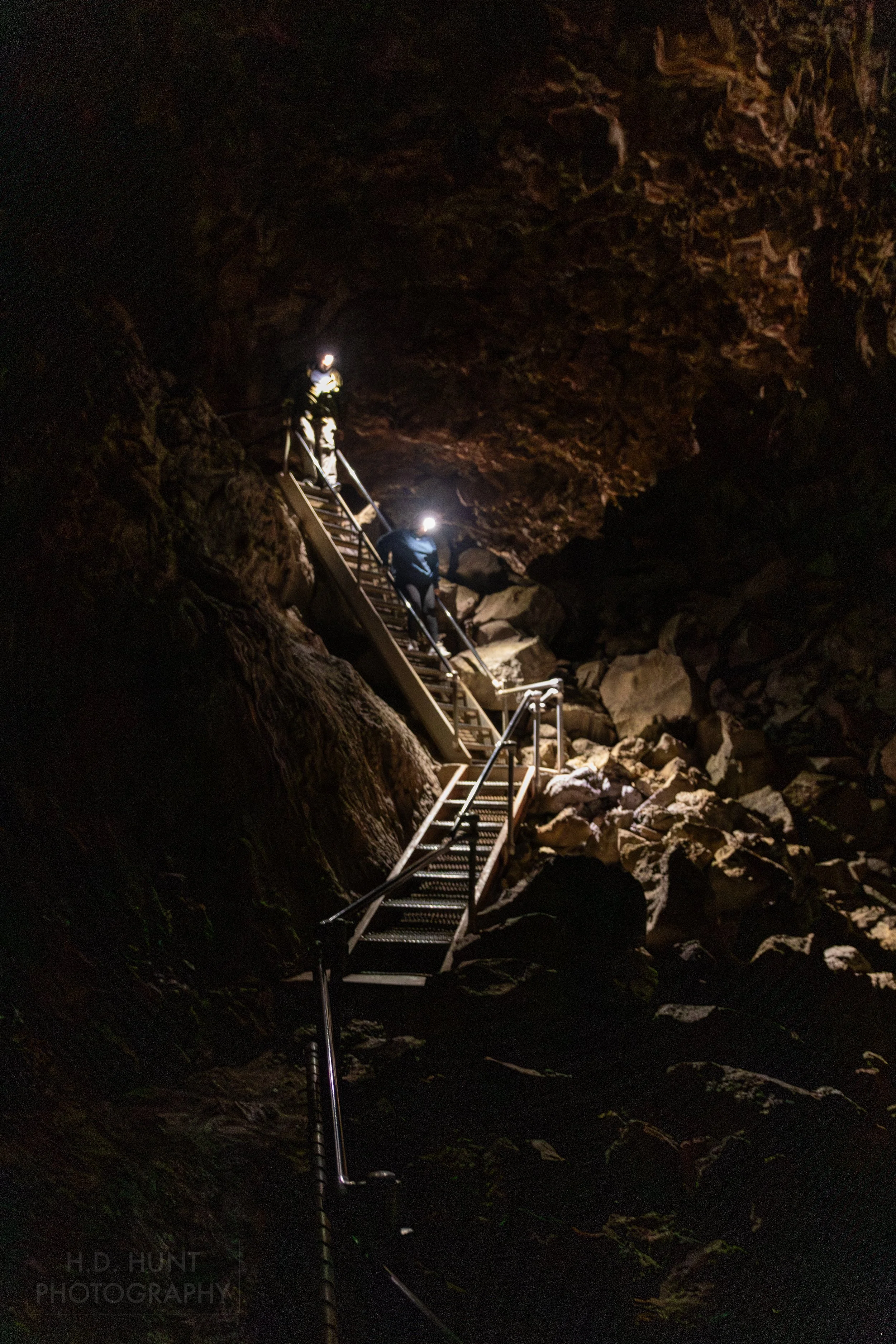 Two visitors descend a steep metal staircase into a dark boulder-covered lava tube, Lava Beds National Monument, California, United States.