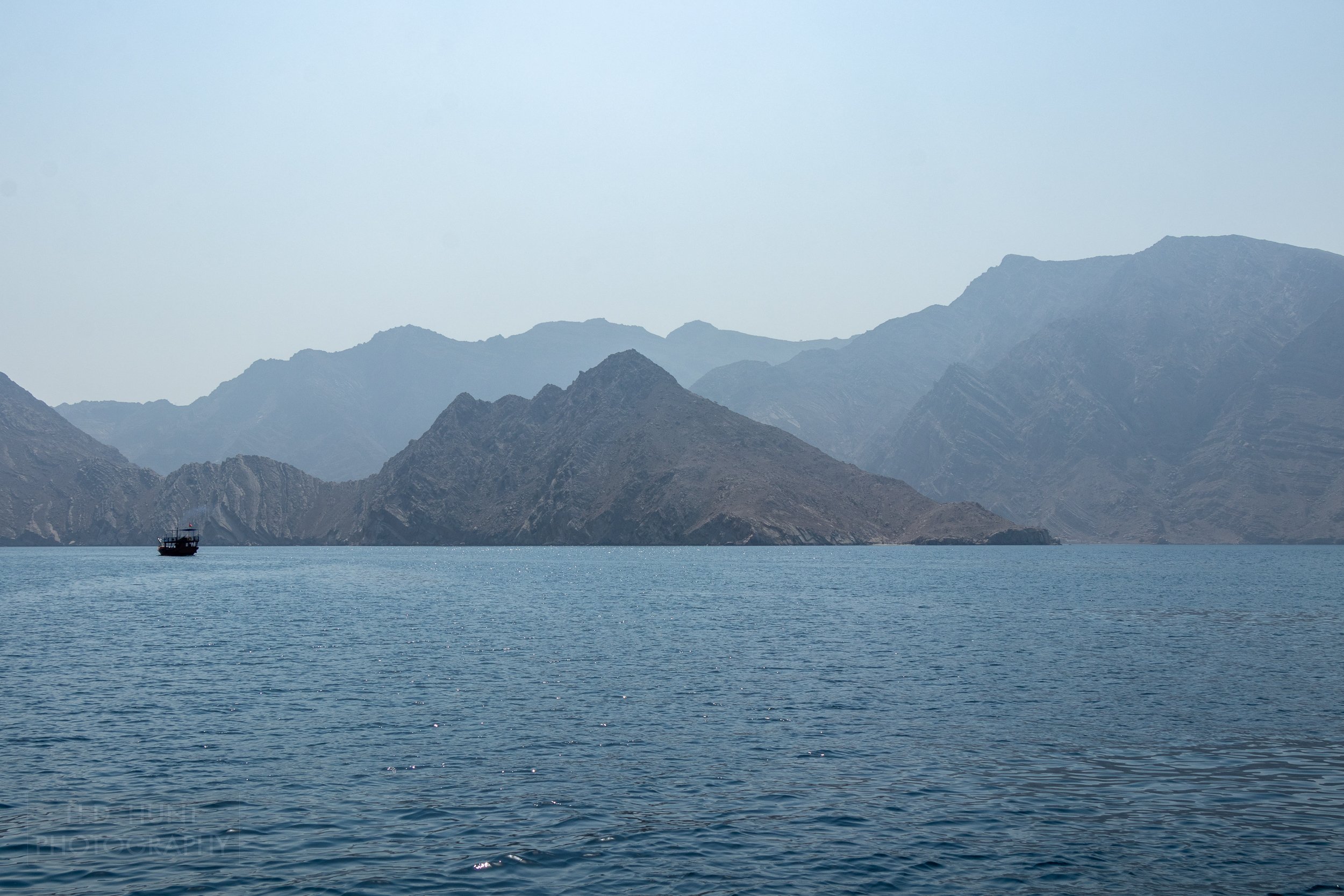A dhow boat is seen in the distance just in front of the mountains of the Musandam Peninsula, Oman.