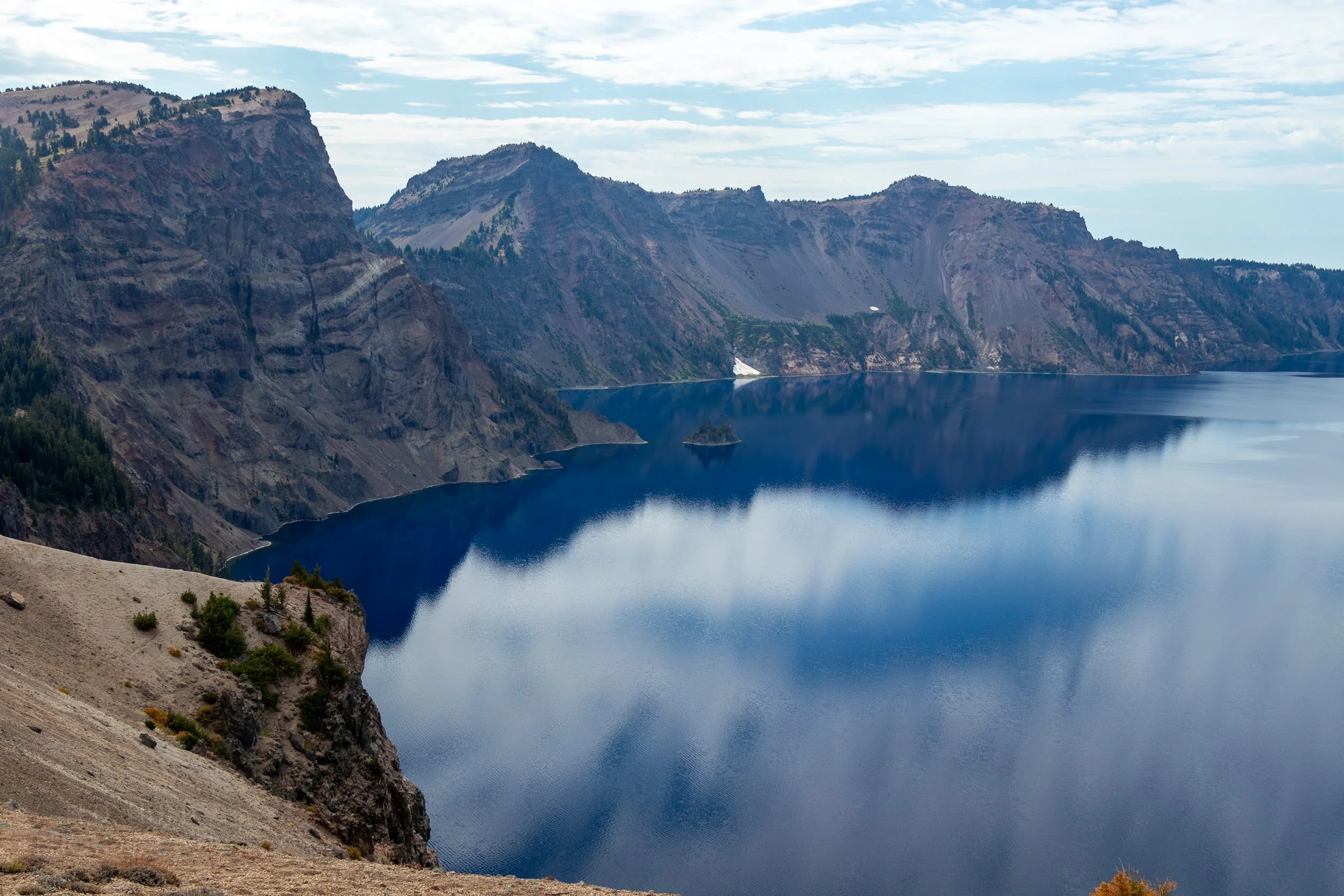The deep blue waters of Crater Lake surrounded by the high cliffs of the Mount Mazama caldera, with the Phantom Ship reaching out above the waterline, Crater Lake National Park, Oregon, United States.
