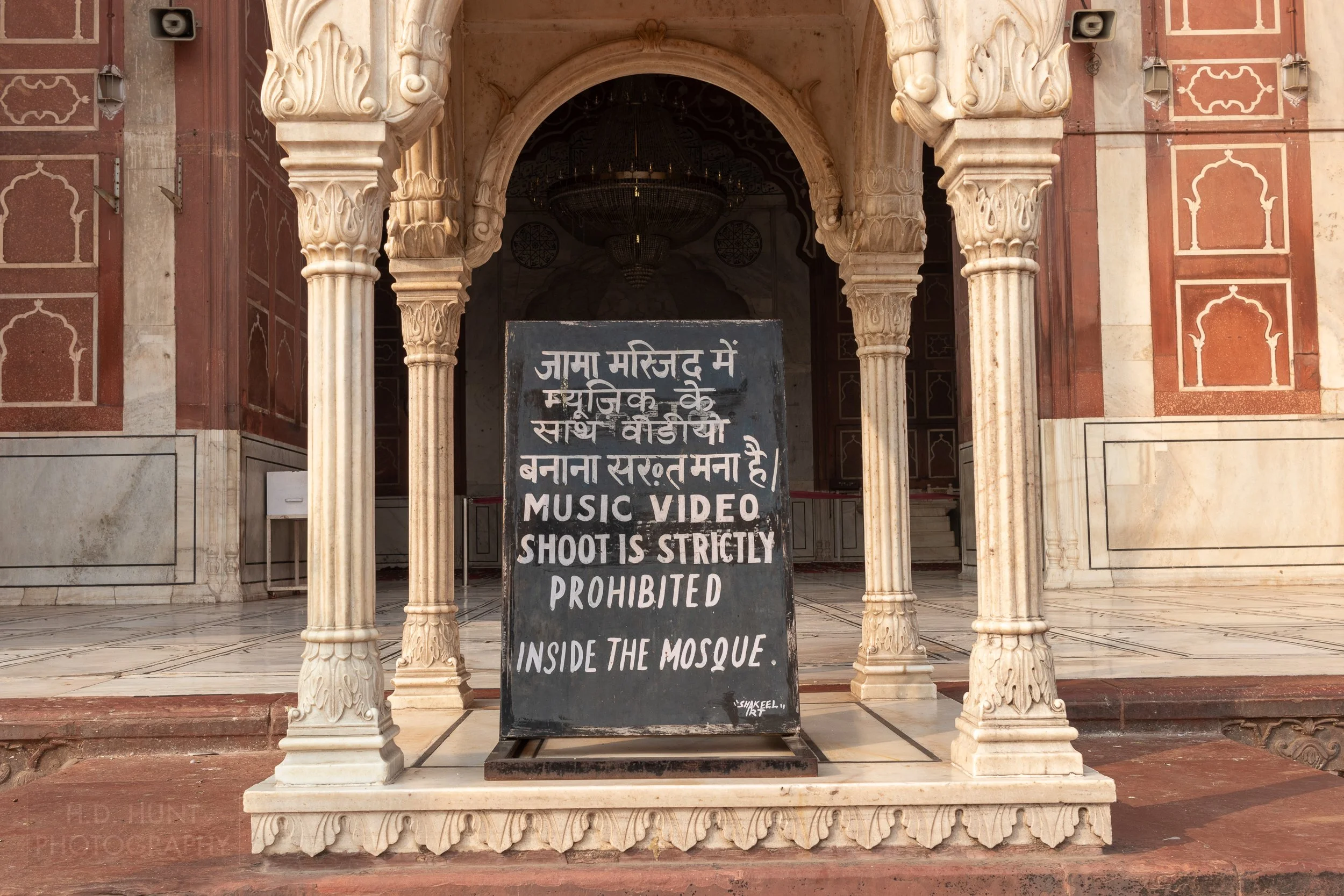 A white-on-black text sign prohibiting music video shooting in the Jama Masjid Mosque, Delhi, India.