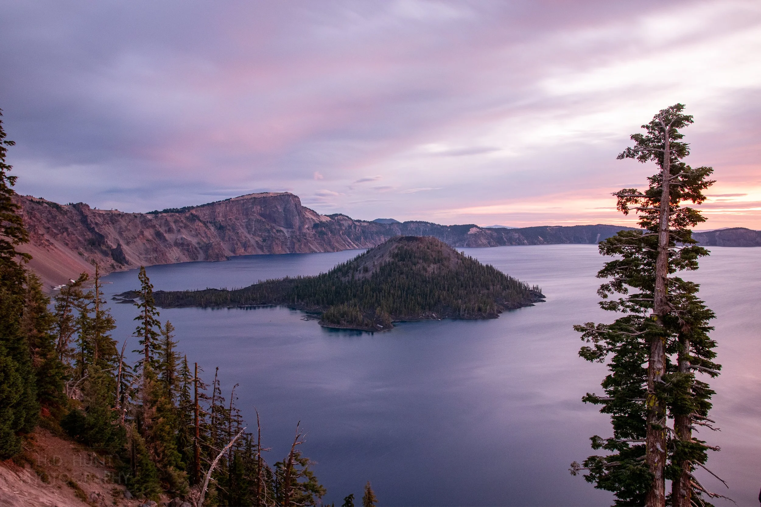 The sunrise creates a pink, orange, and yellow sky above the blue waters of Crater Lake and Wizard Island, Crater Lake National Park, Oregon, United States.