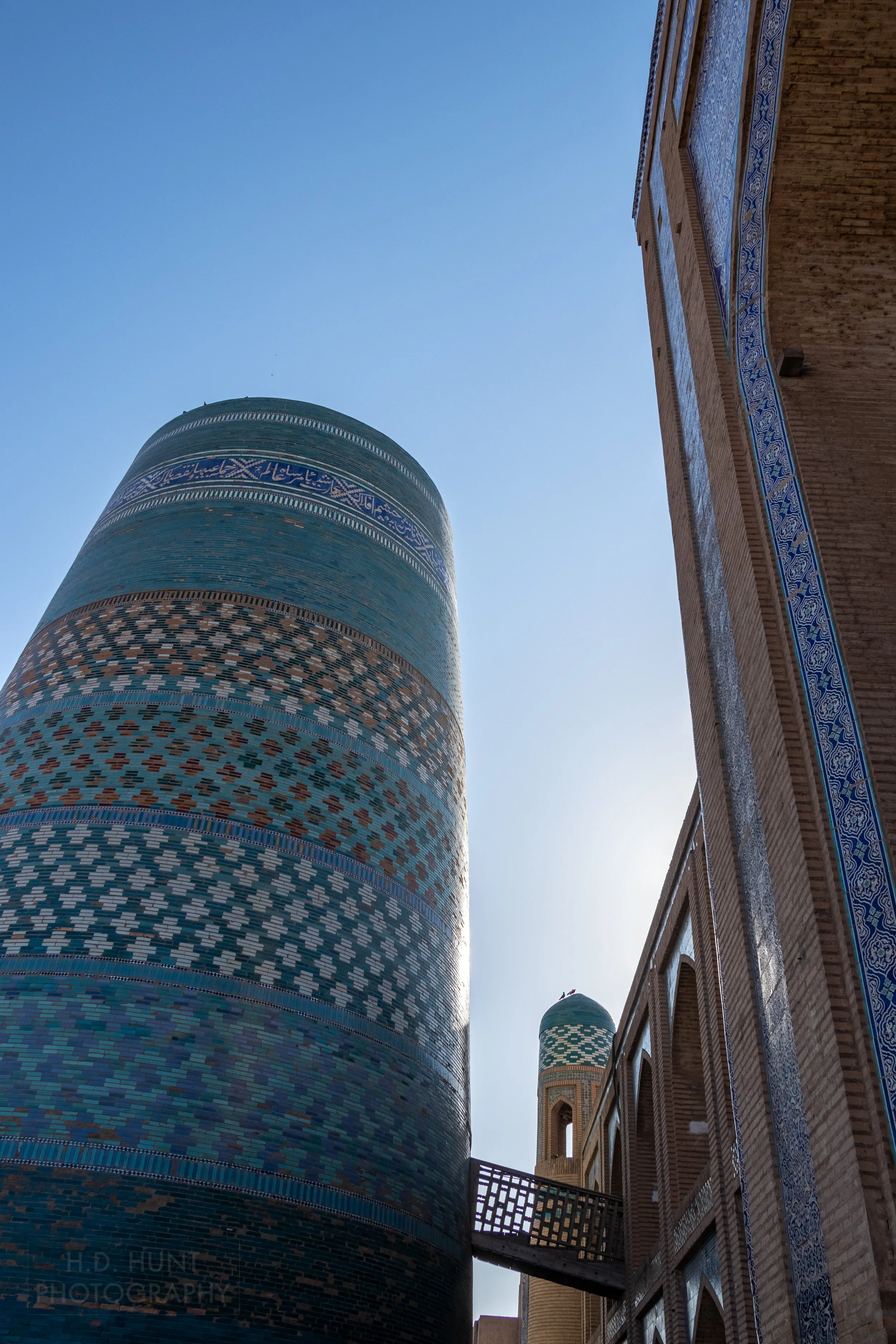 A wooden walkway connects the tiled Kalta Minor tower and the brick Muhammad Amin Khan Madrasa in Khiva, Uzbekistan.