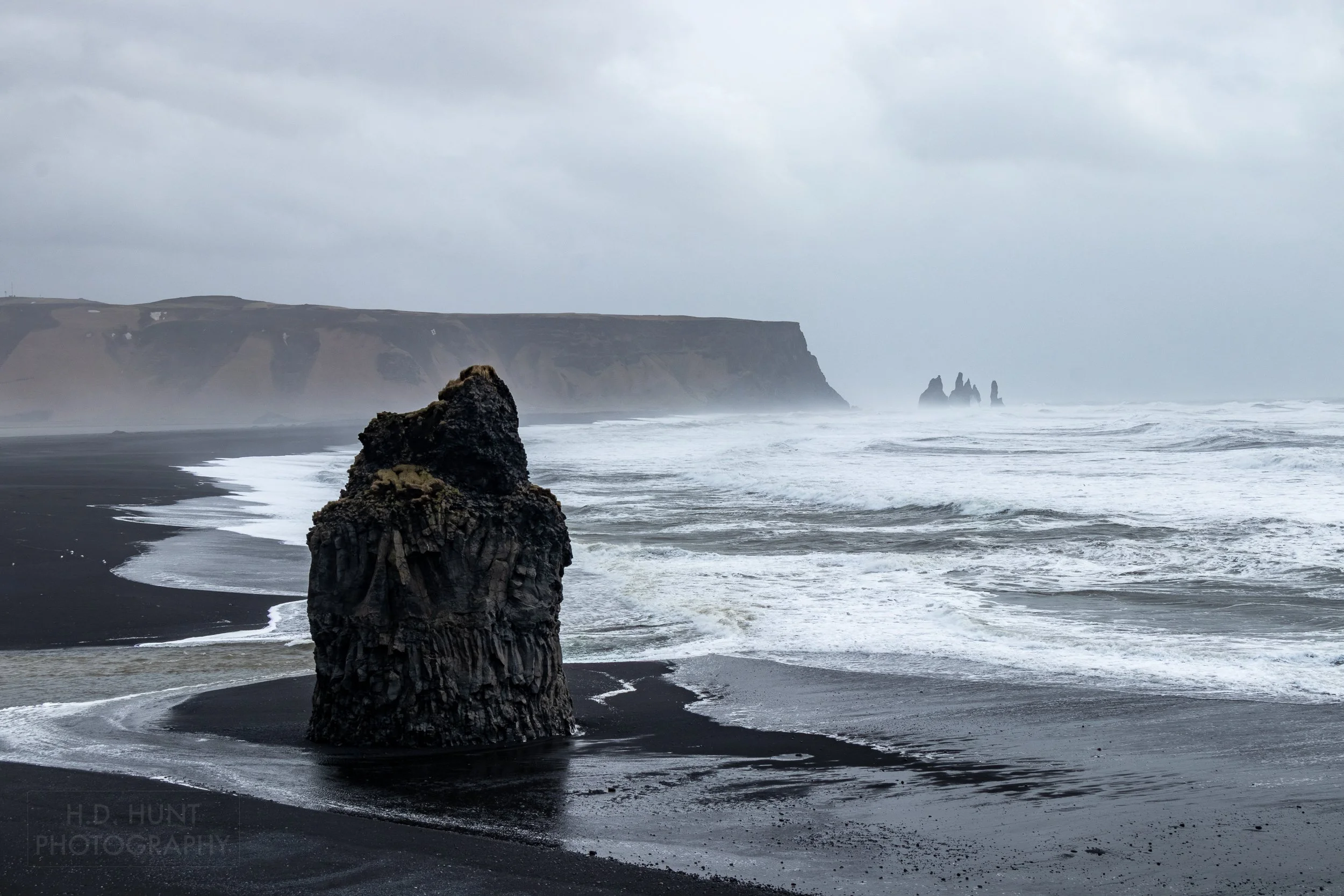 A large basalt column rises above a black sand beach beach which stretches into the background before meeting a tall, flat cliff, Dyrhólaey, Iceland.