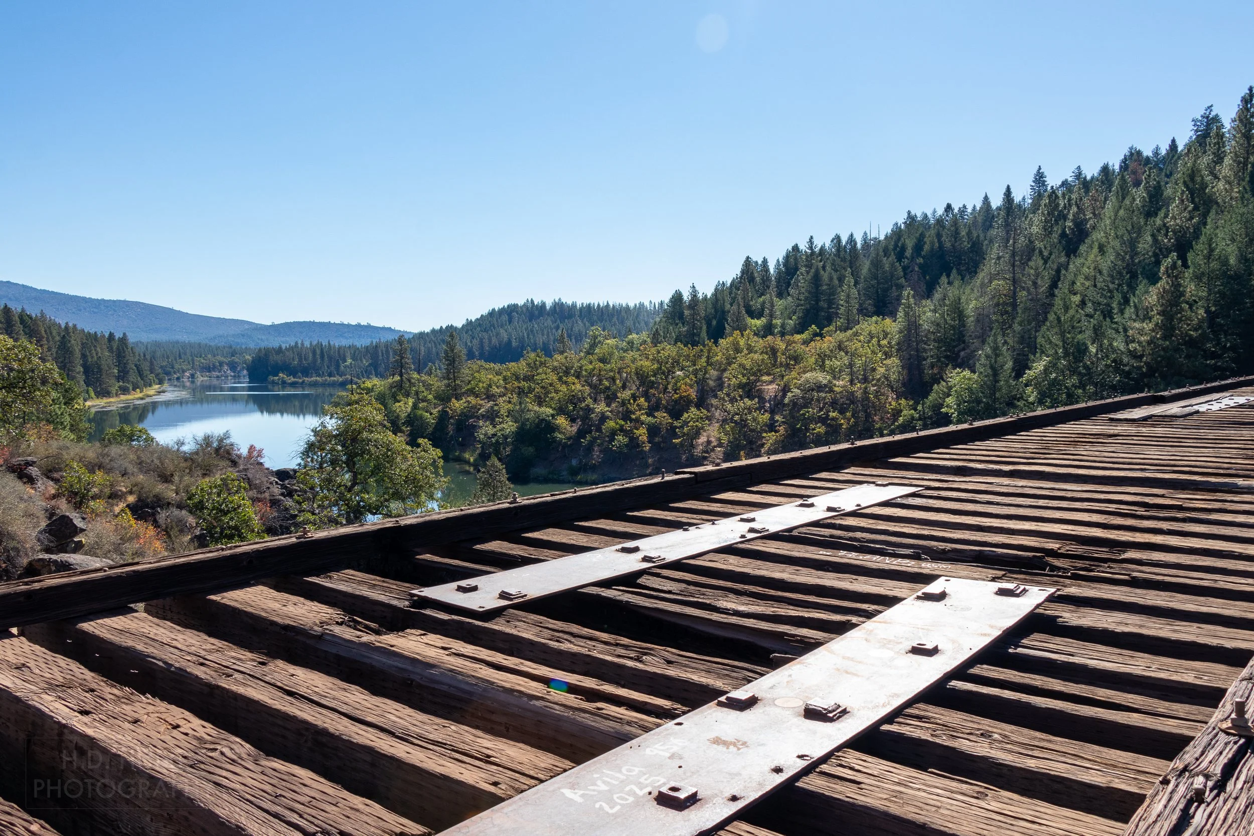 A railroad trestle bridge with brown timbers but no tracks spans a green river before disappearing behind trees, California, United States.