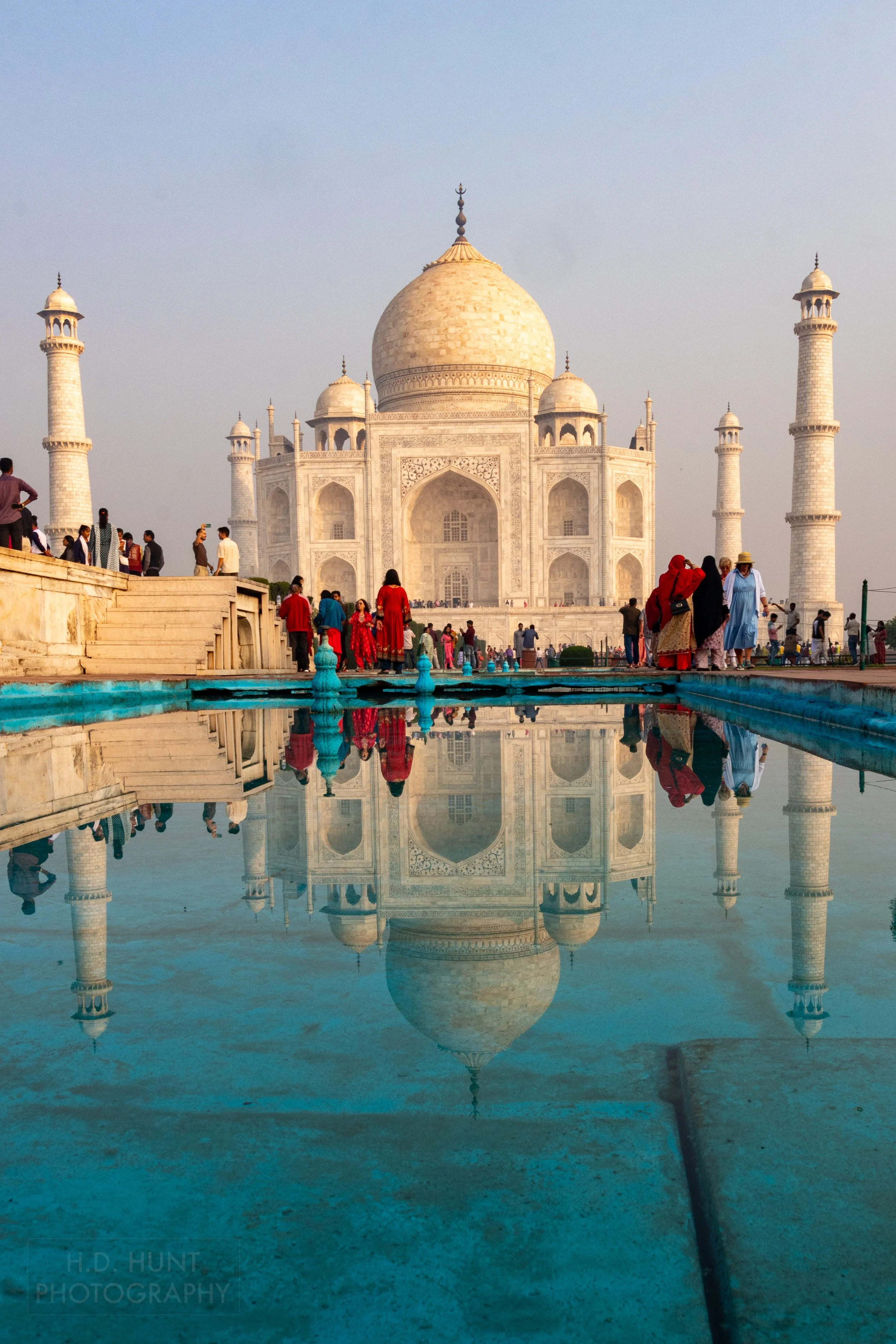 Several visitors - many dressed in bright red clothing - stand between the Taj Mahal and a blue reflecting pool, Agra, India.