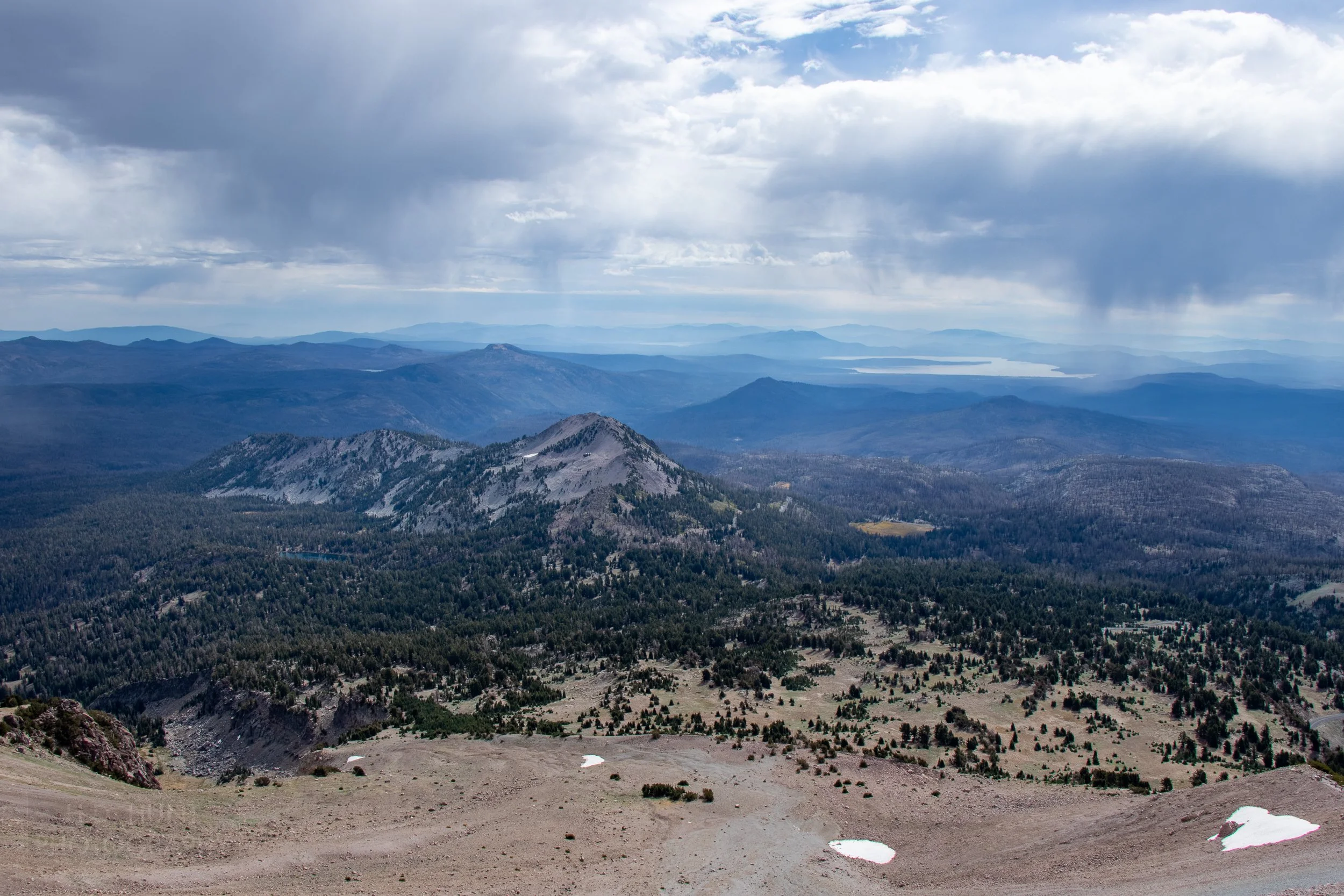 Trees and mountains stretch into the distance towards a large lake underneath thick grey clouds, as seen from Lassen Peak, Lassen Volcanic National Park, California, United States.