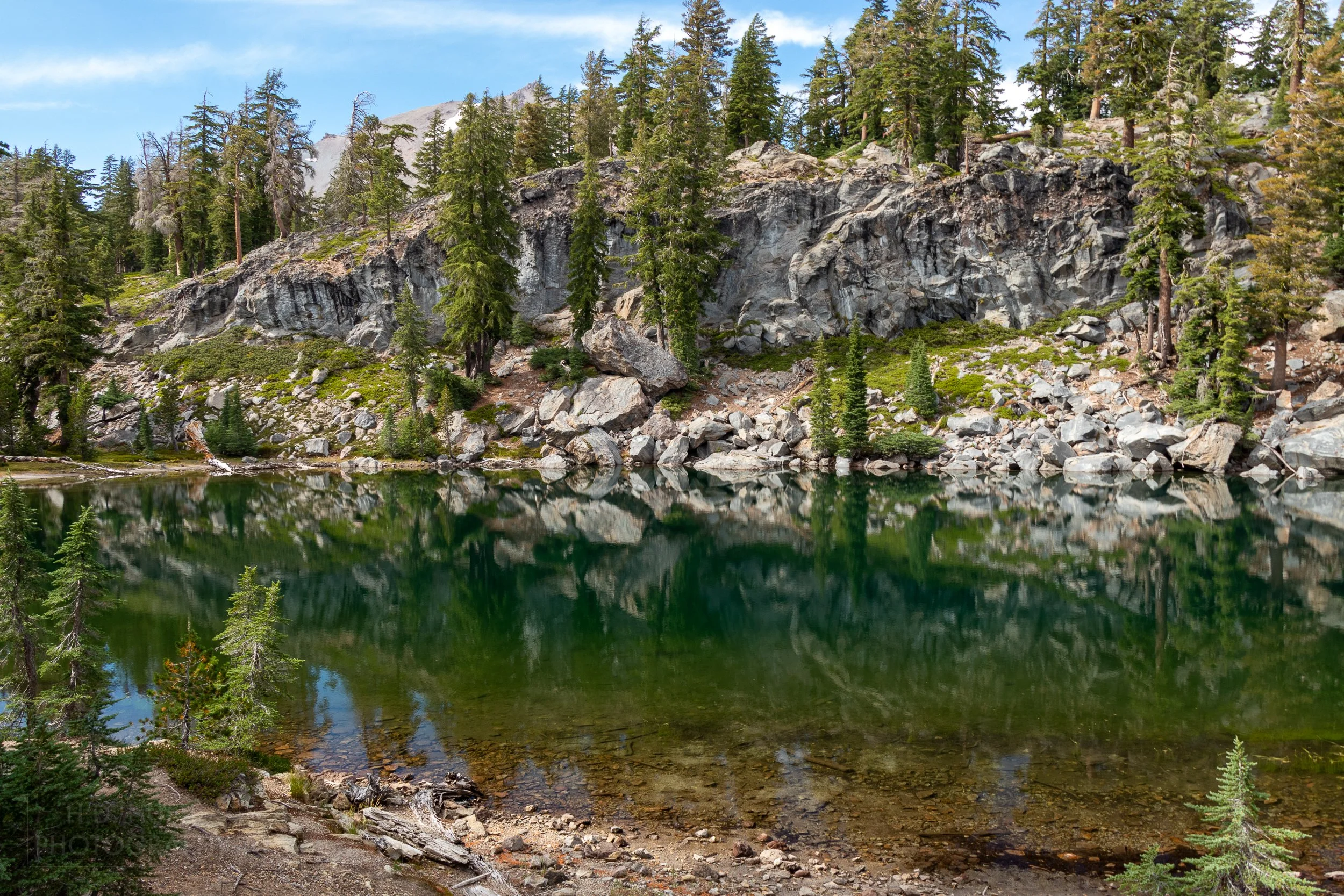 A green lake reflects a nearby rock cliff and trees, Lassen Volcanic National Park, California, United States.