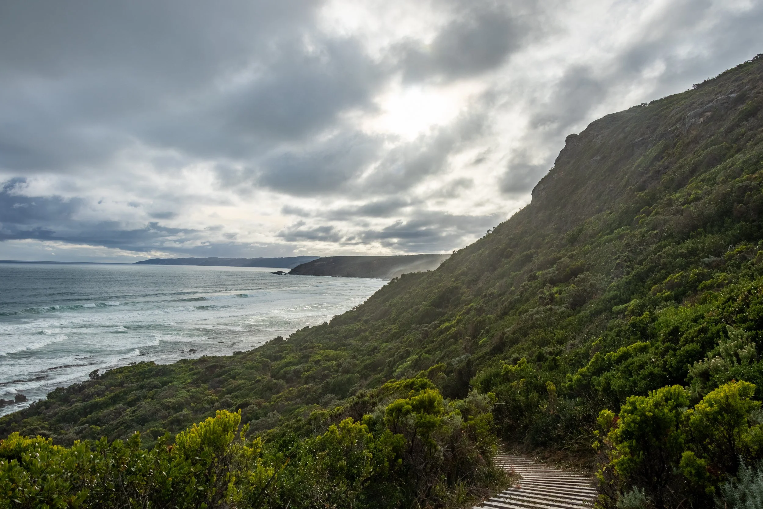 A wooden staircase descends towards the Southern Ocean beneath a tall green shrub-covered cliff along The Great Ocean Walk, Victoria, Australia.