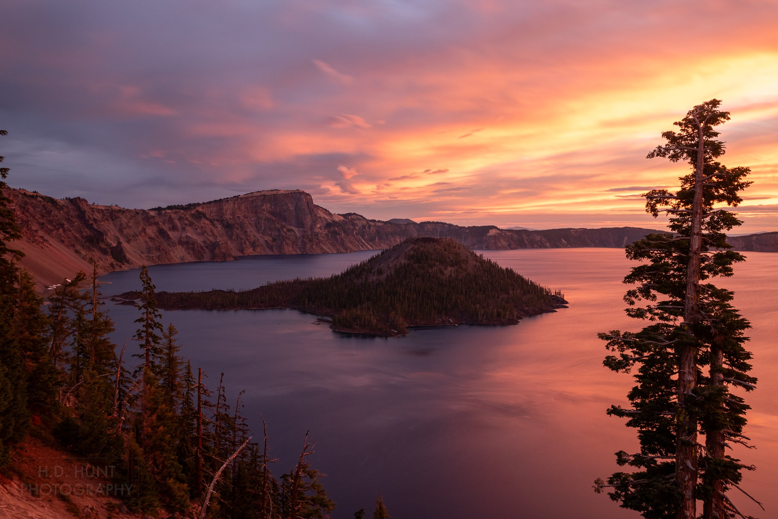 The sunrise creates a pink, orange, and yellow sky above the blue waters of Crater Lake and Wizard Island, Crater Lake National Park, Oregon, United States.