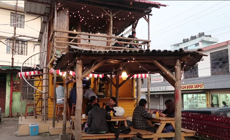 Outdoor café in Shillong reflecting the city’s thrift culture, with bamboo structure, string lights, and young people dining at wooden tables.