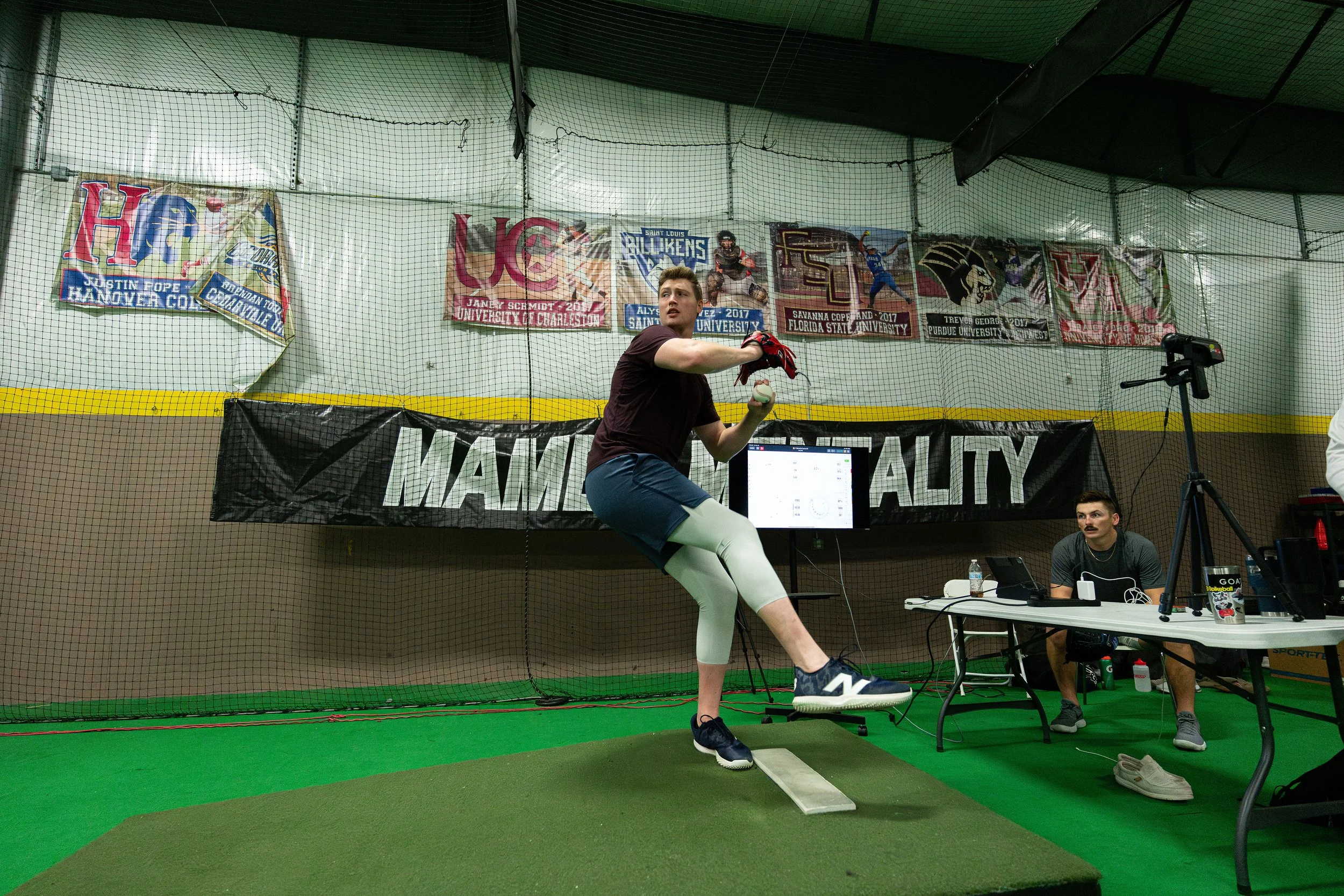A person pitching a baseball in an indoor facility with banners hanging on the wall behind. The person is wearing a black shirt, blue shorts, white leggings, and black sneakers. There is a person sitting at a desk with computer equipment nearby.