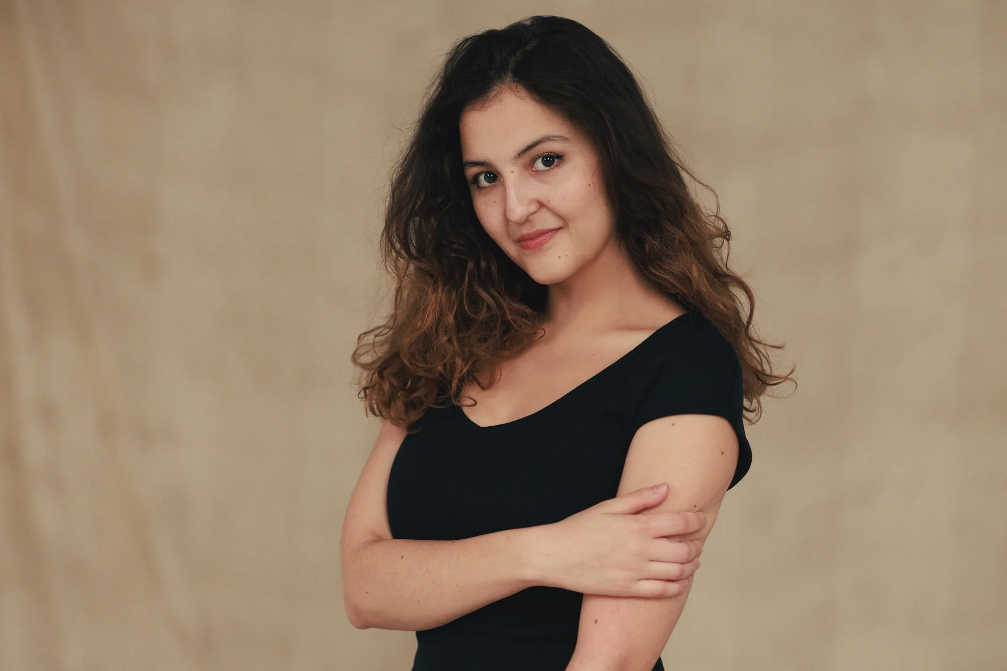 Young woman with long curly brown hair, wearing a black top, standing against a beige background, smiling softly with arms crossed.