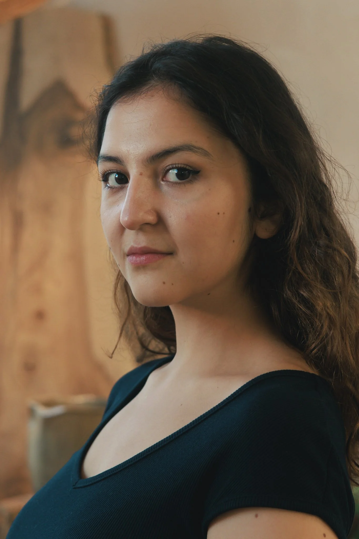A woman with curly brown hair and light skin, wearing a black top, looking at the camera with a subtle smile, with warm indoor lighting and a blurred wooden background.