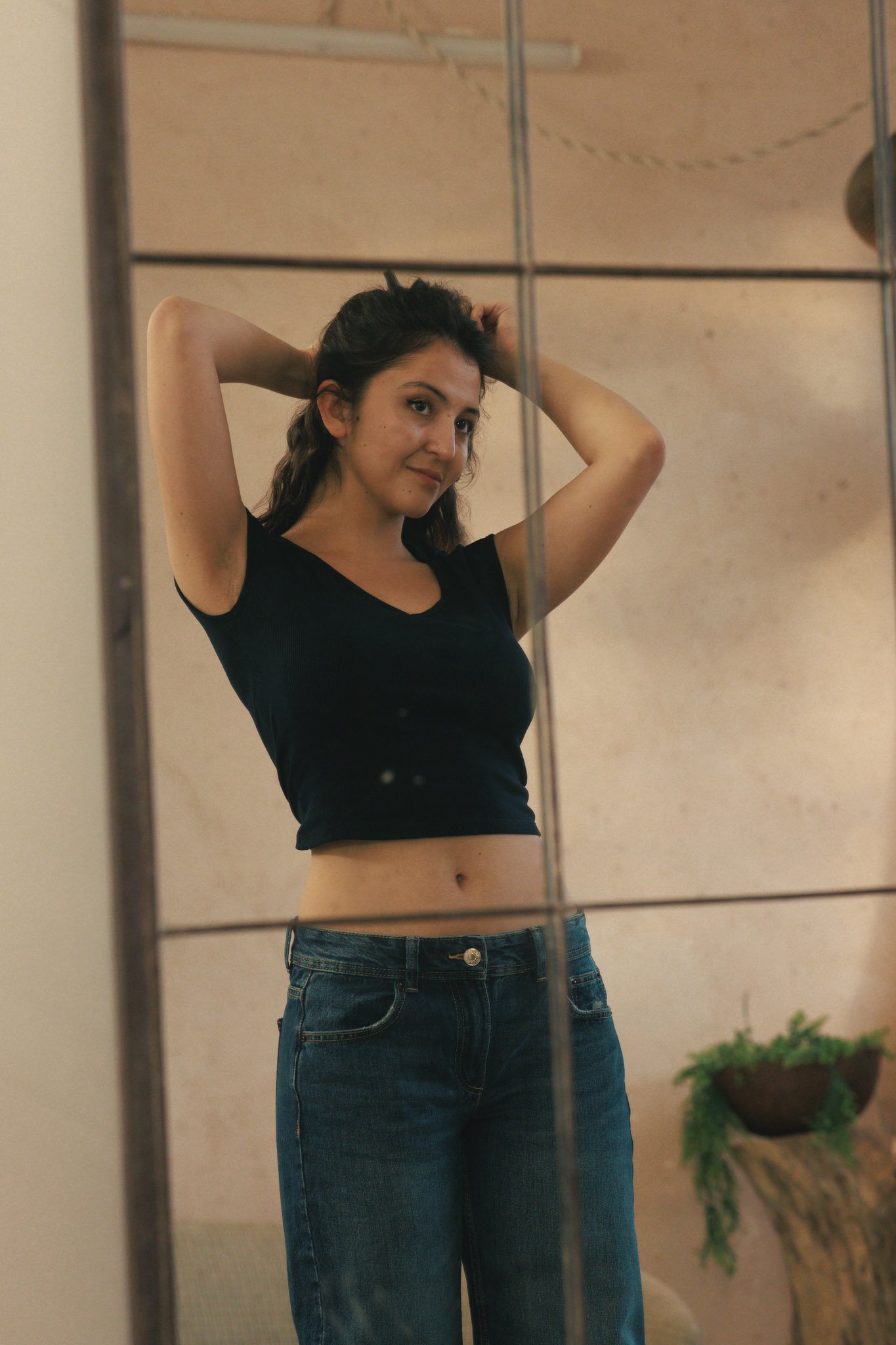 A young woman with long brown hair wearing a black crop top and blue jeans, standing indoors in front of a mirror, adjusting her hair.
