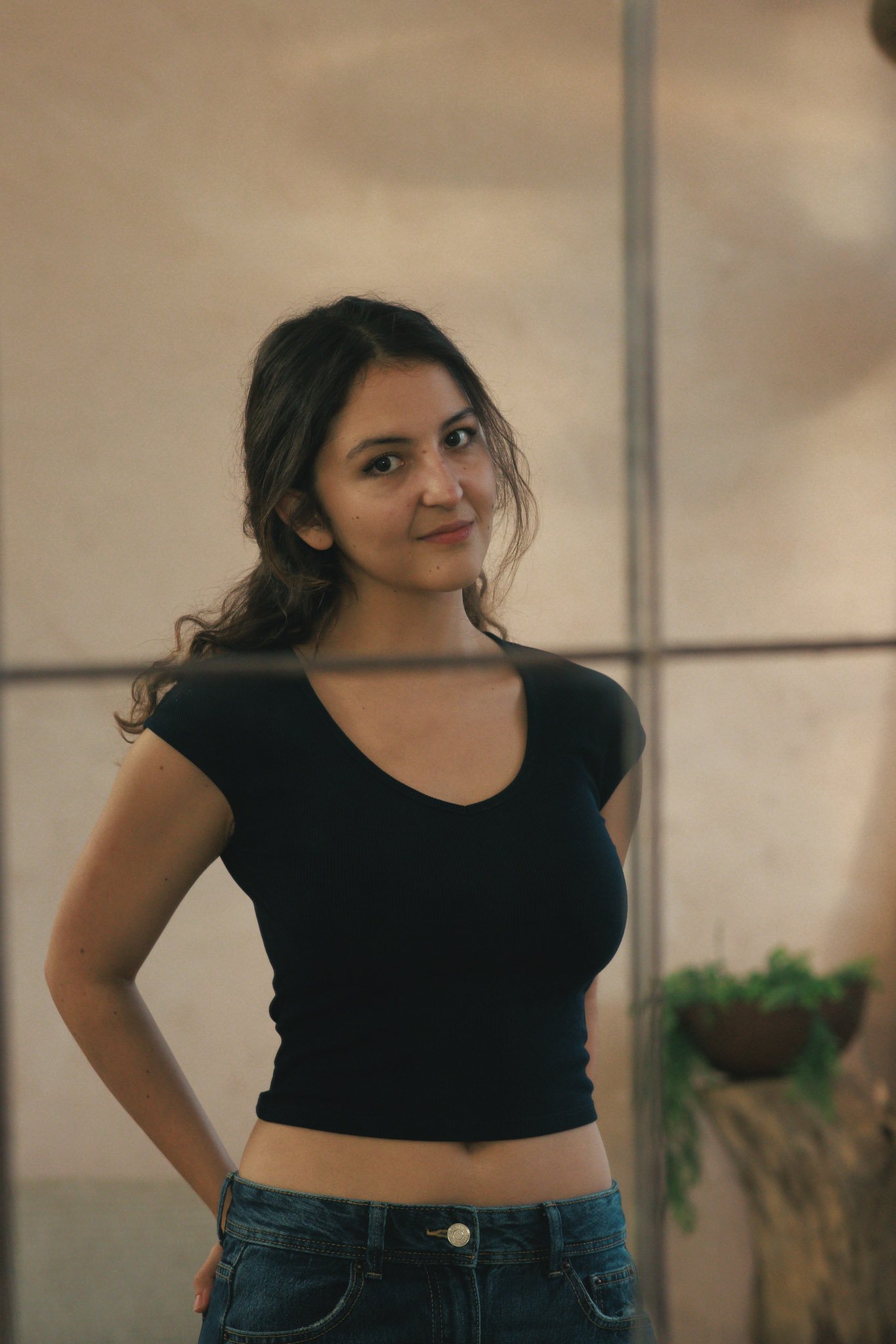 A young woman with long brown hair wearing a black crop top and blue jeans, standing indoors with a neutral expression.