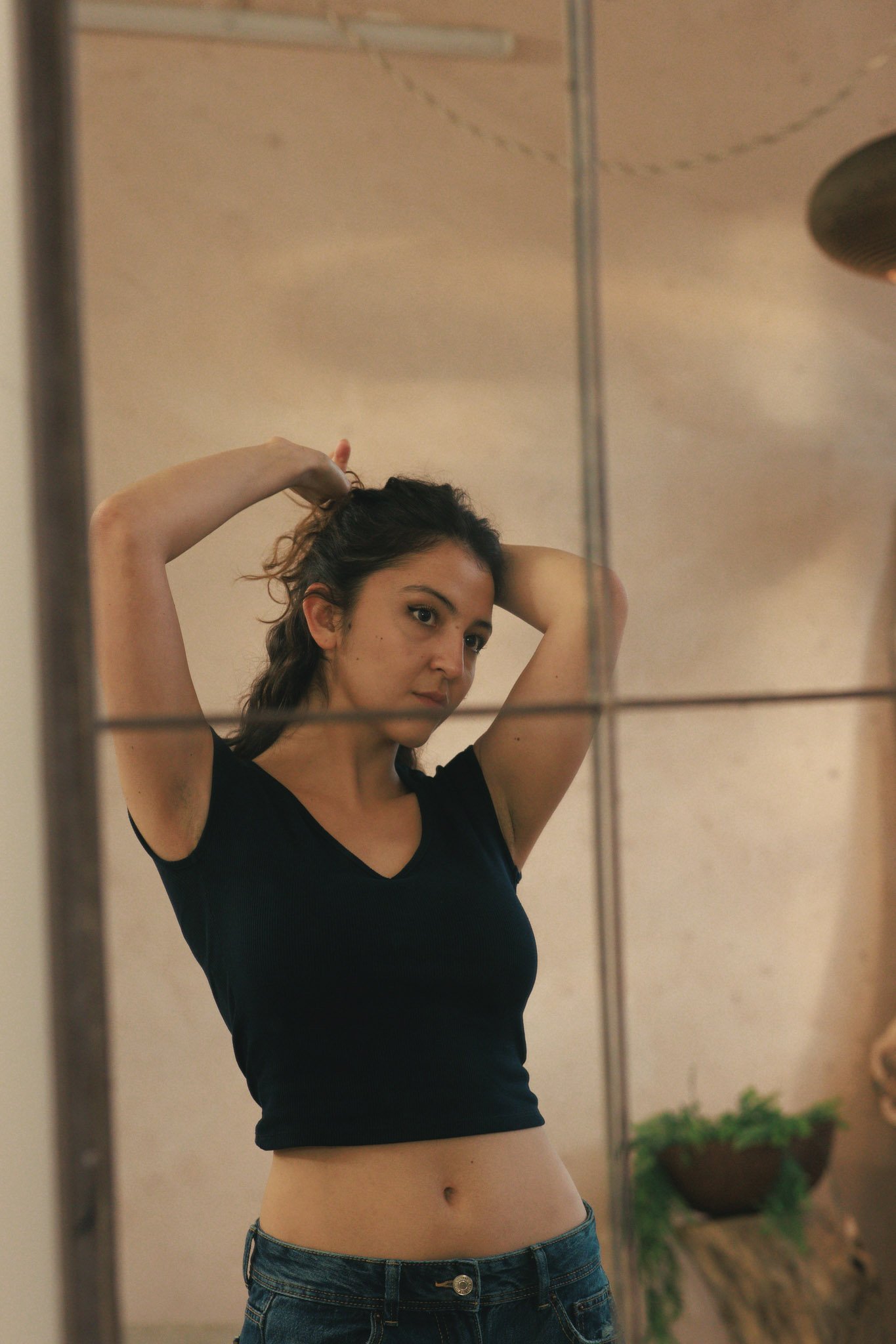 Young woman taking a mirror selfie, wearing a black crop top and blue jeans, with her hands in her hair.