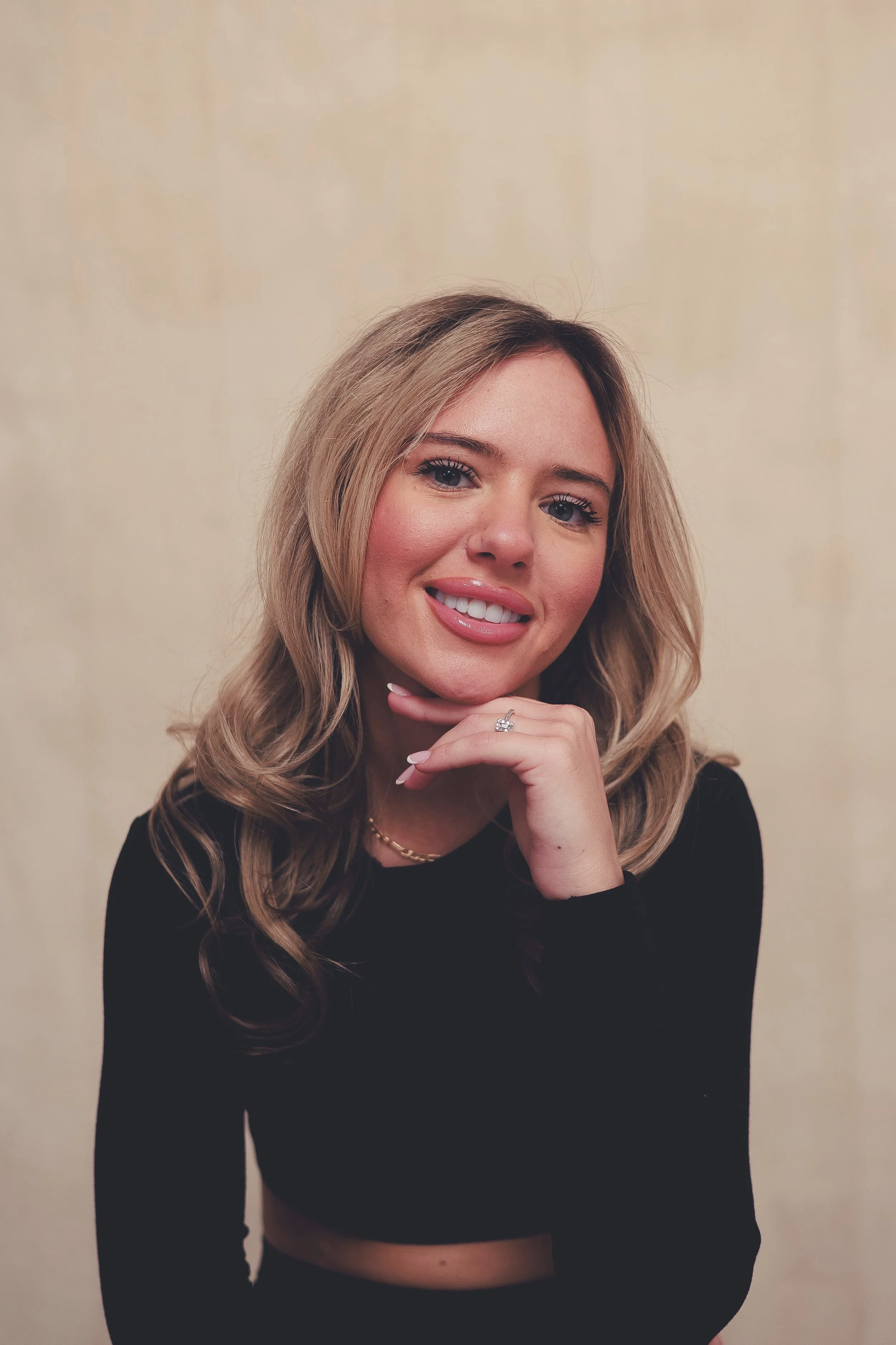 A young woman with blonde hair and blue eyes, smiling, resting her chin on her hand, wearing a black top and jewelry, against a plain beige background.