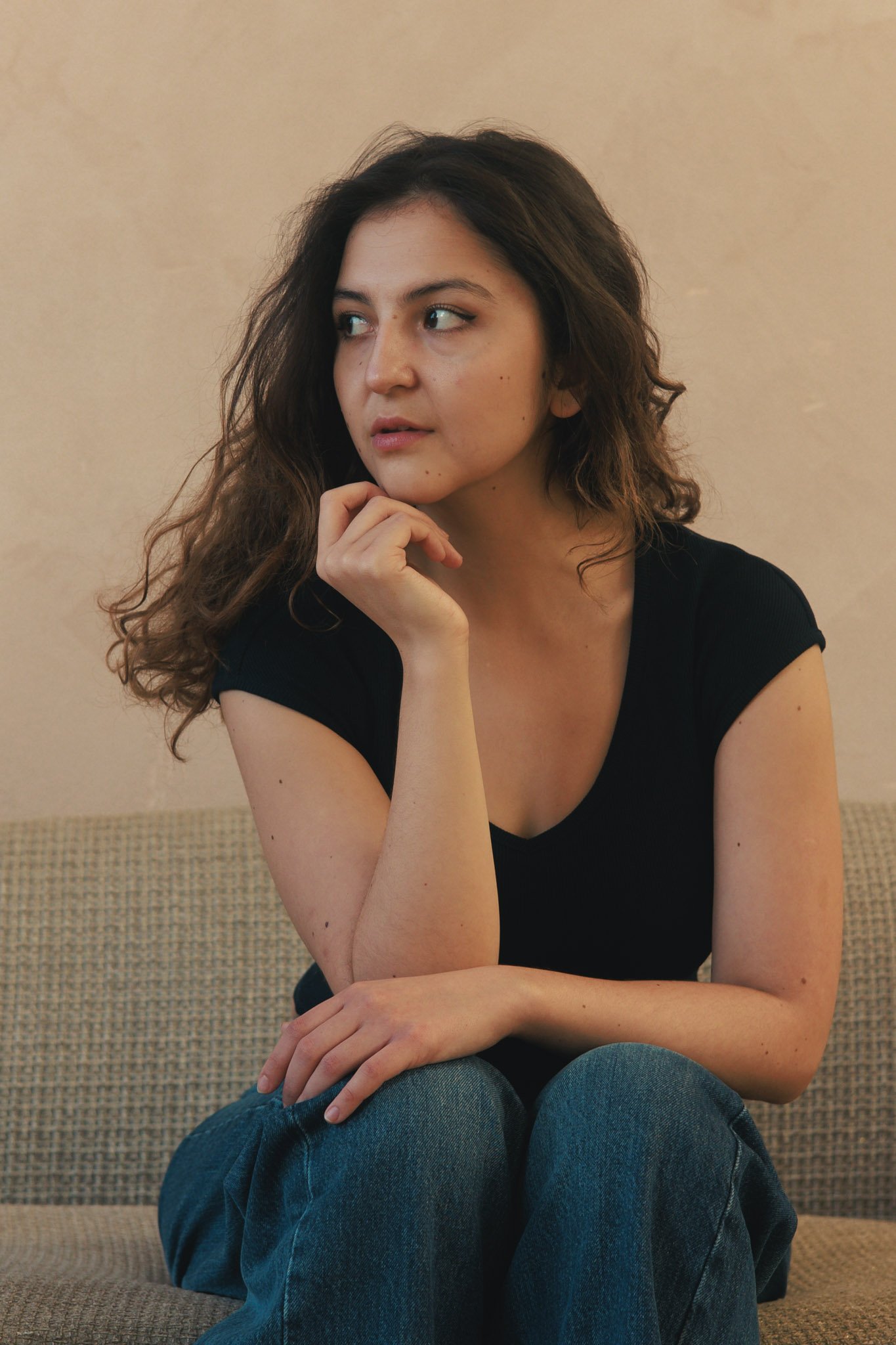 A young woman with wavy brown hair sitting on a beige couch, wearing a black t-shirt and blue jeans, resting her chin on her hand, looking to her left with a thoughtful expression.